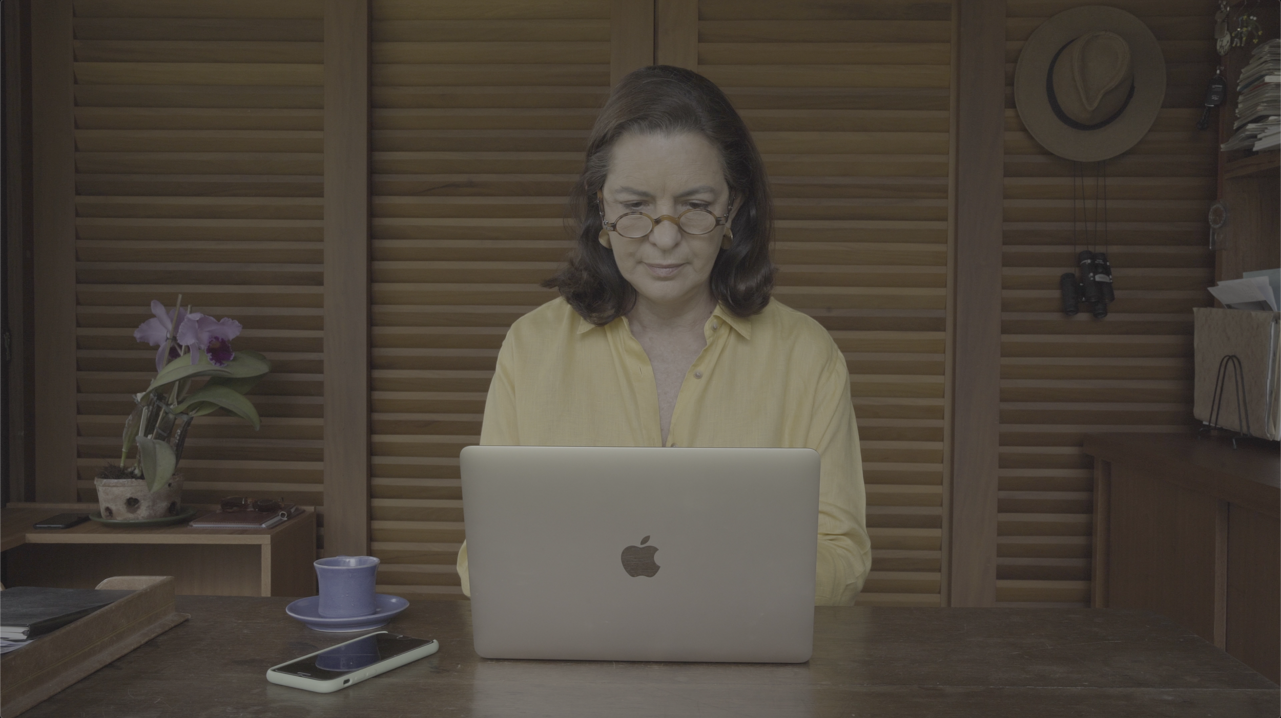 A woman with glasses and dark hair looking at a silver MacBook laptop on a wooden desk in a room with wooden panel walls, a hat hanging on the wall, and a purple orchid plant on a side table.