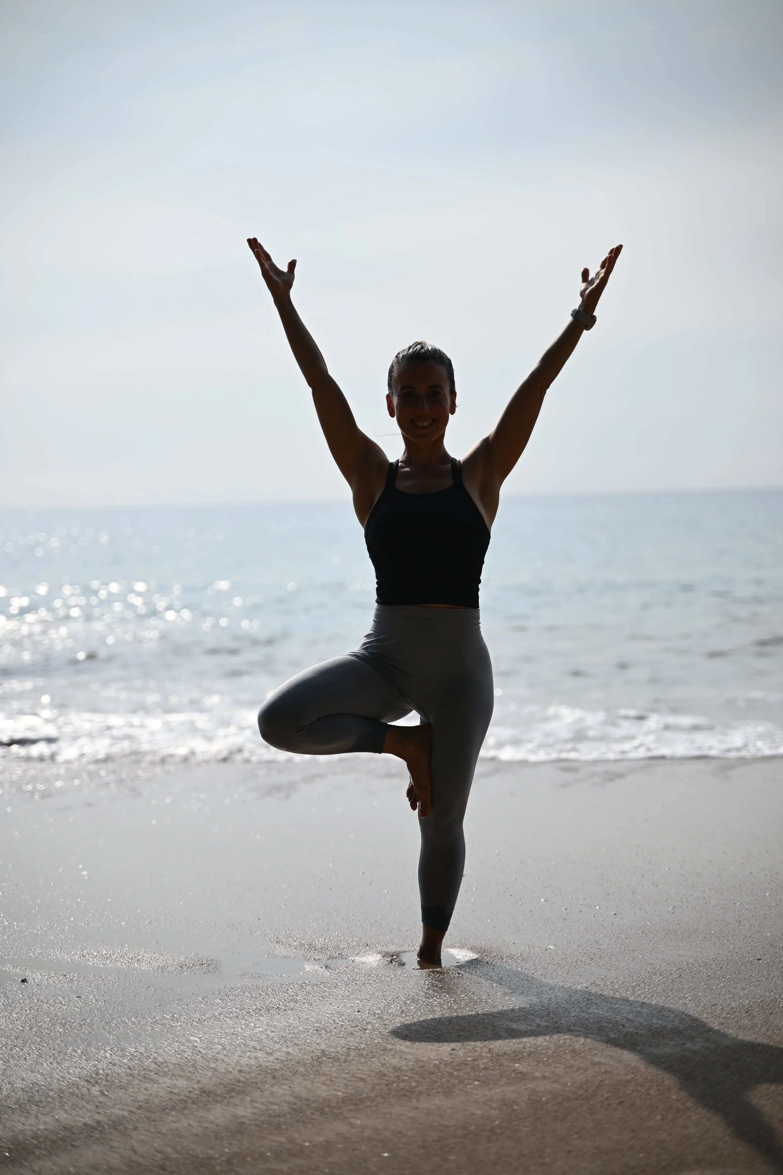 A woman practicing yoga on the beach, performing a tree pose with arms raised, near the shoreline with the ocean in the background.