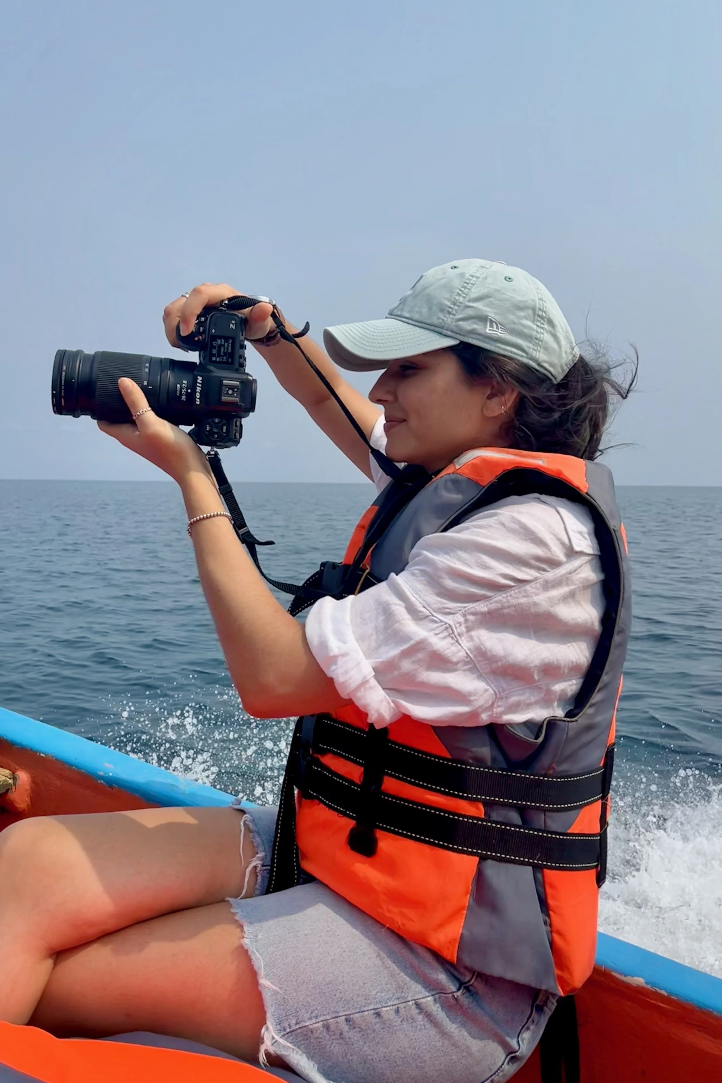 Woman wearing a gray cap and orange life jacket, sitting on a boat, taking a photograph with a DSLR camera by the ocean under a hazy sky.
