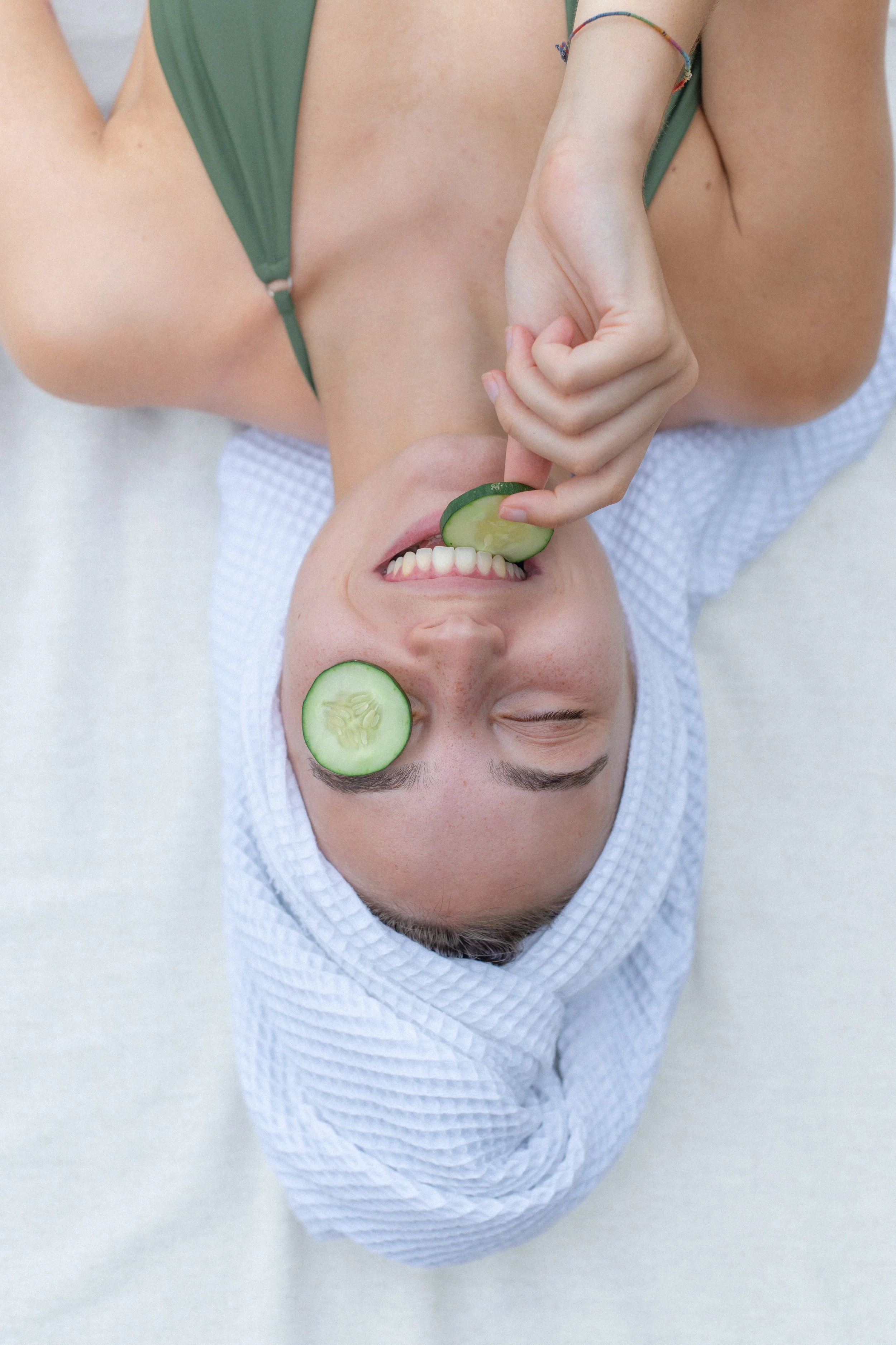 A woman lying down with her head on a white pillow, wearing a green top and a white towel wrapped around her head. She is smiling, with a slice of cucumber over one eye, and holding a cucumber piece near her mouth.