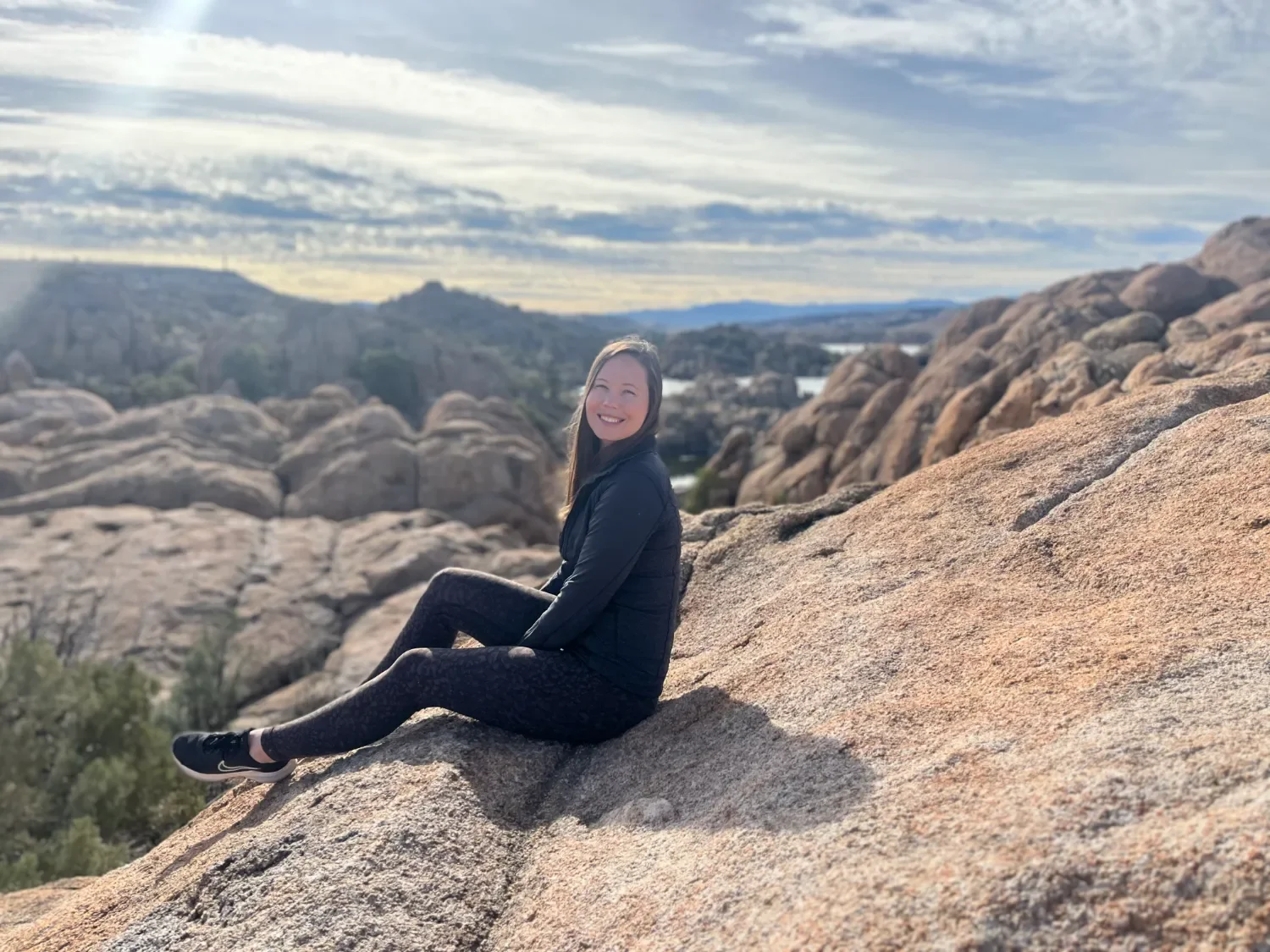 Emily Sarff Summit Somatics sitting on a large rock formation outdoors, smiling with a scenic landscape of rocks, trees, and cloudy sky in the background.