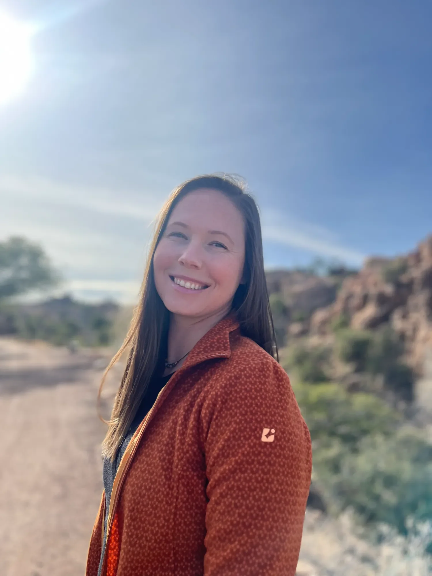 Emily Sarff Summit Somatics smiling outdoors in a desert landscape with rocky hills and sparse vegetation.