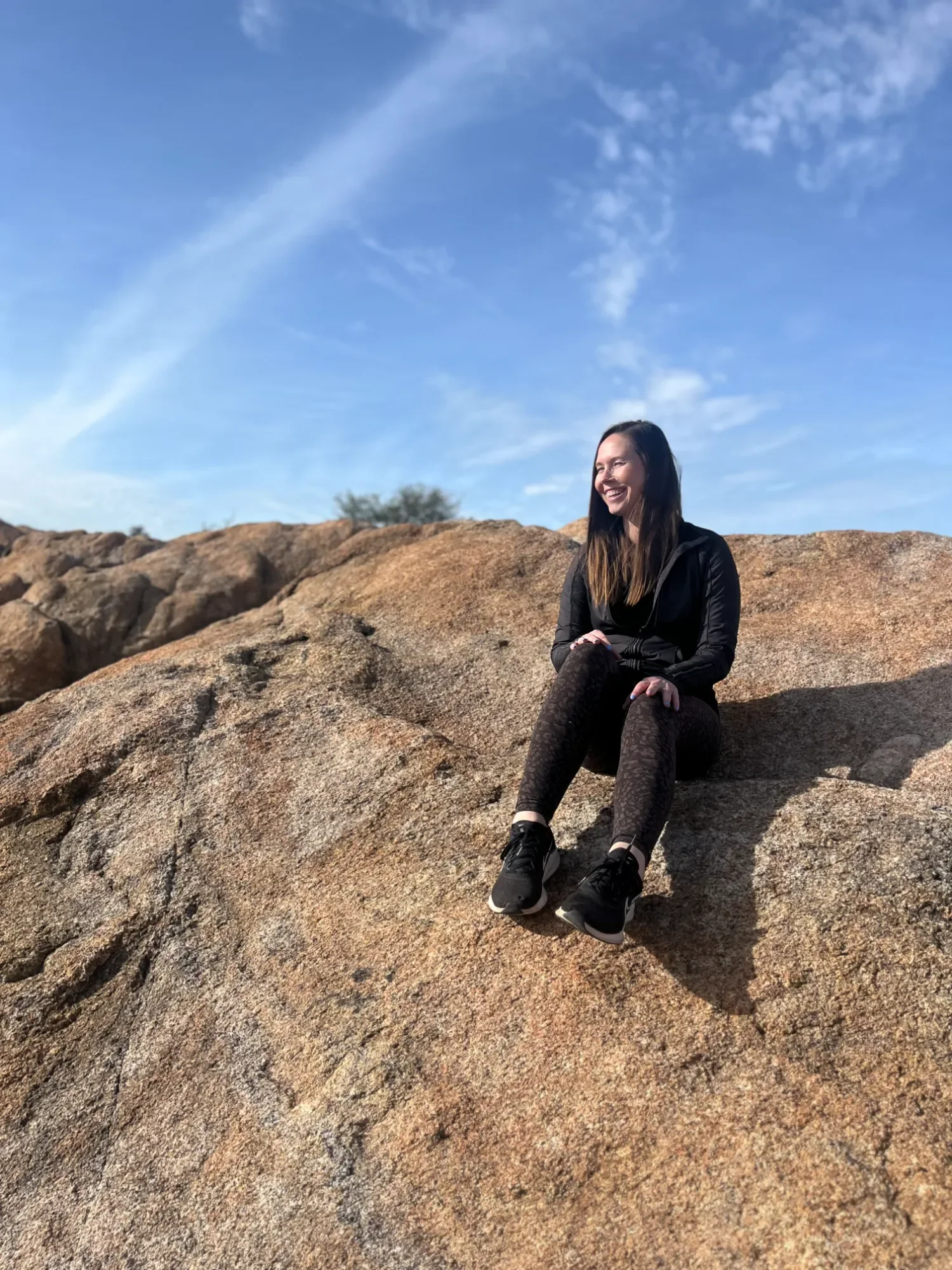 Emily Sarff Summit Somatics sitting on a large rock outdoors with a bright blue sky and wispy clouds behind her, smiling.