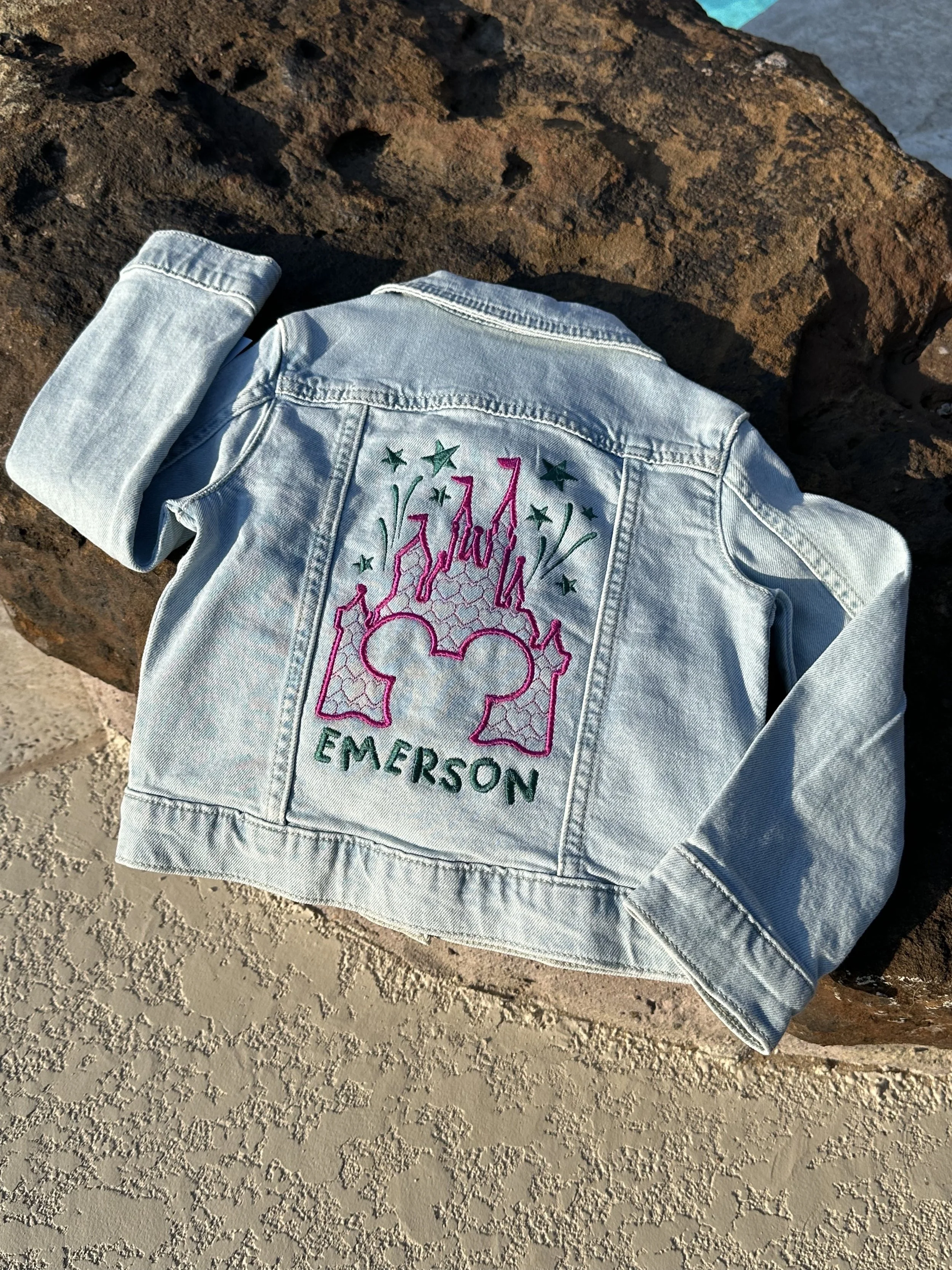 Light blue denim jacket with colorful embroidery of fireworks, stars, a castle silhouette, and the name 'Emerson' on the back, placed on a large rock.