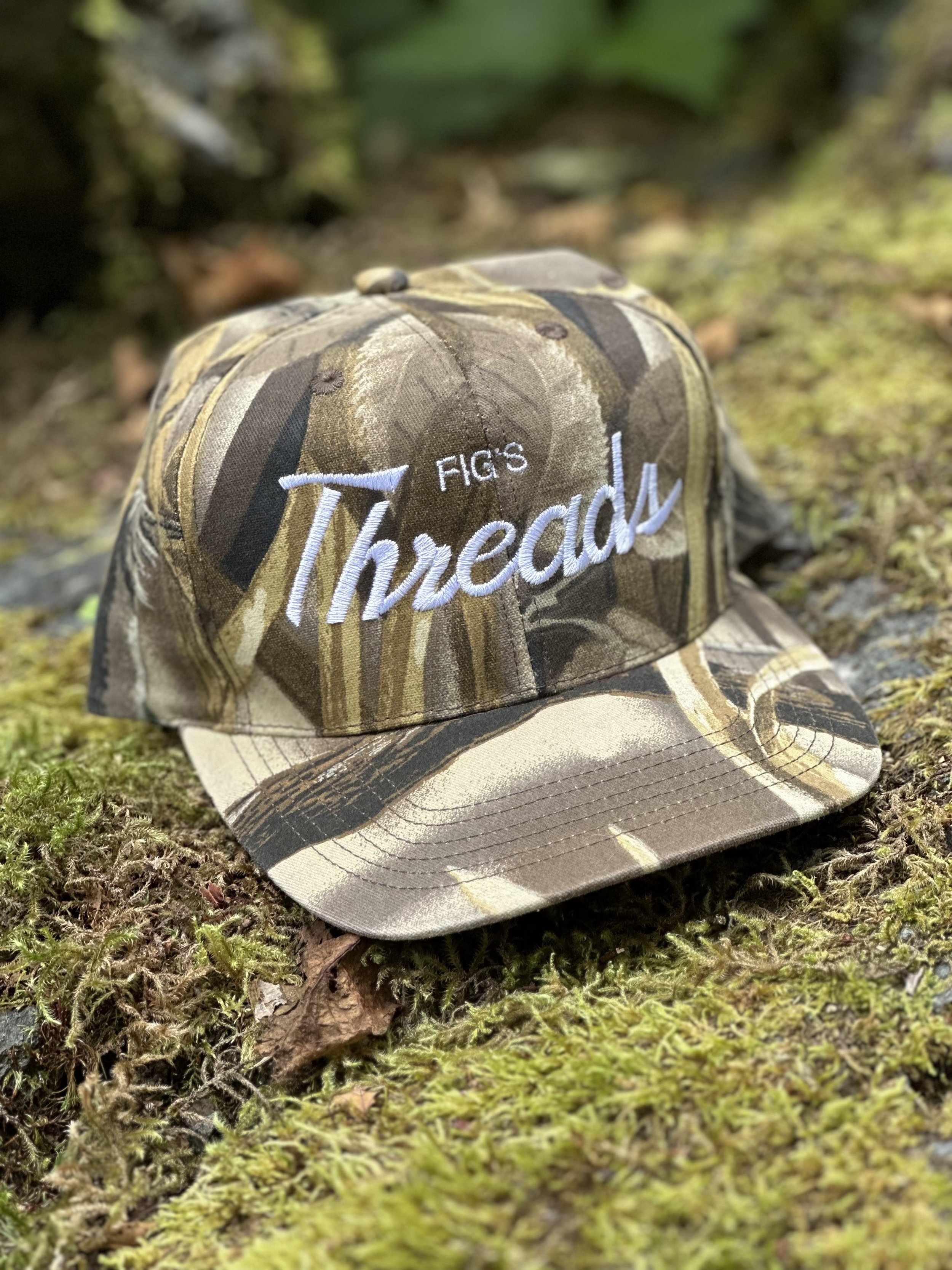 Camouflage cap with white embroidered text 'Fig's Threash' resting on mossy ground in a forest