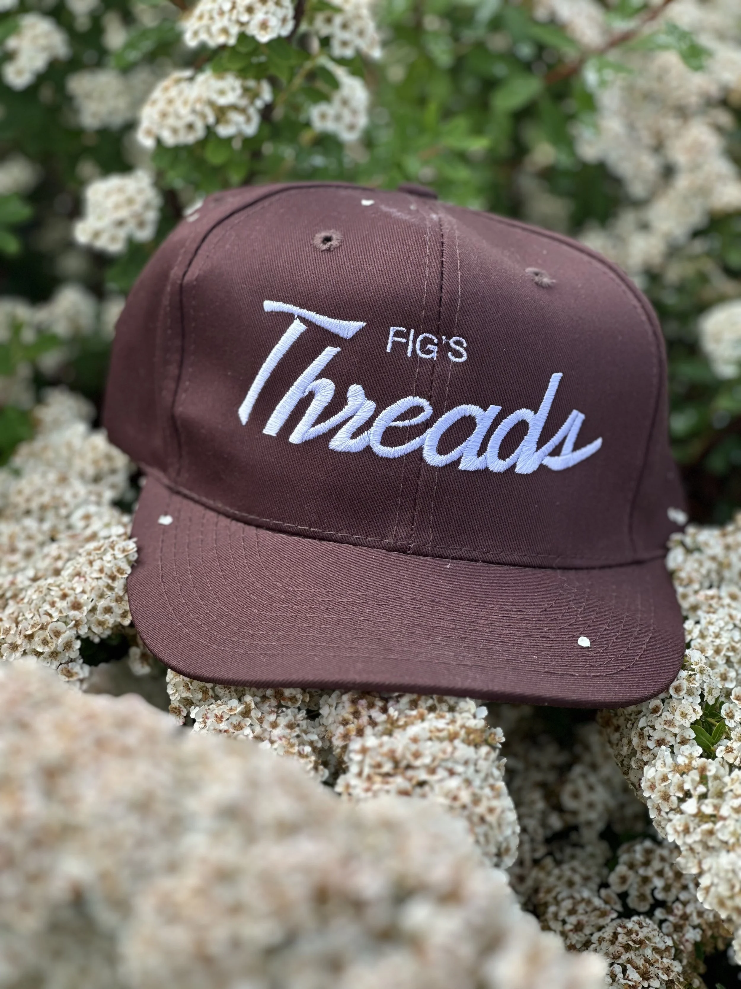 A maroon baseball cap with white embroidered text reading 'FIG'S Threads' placed on white flowers with green leaves in the background.