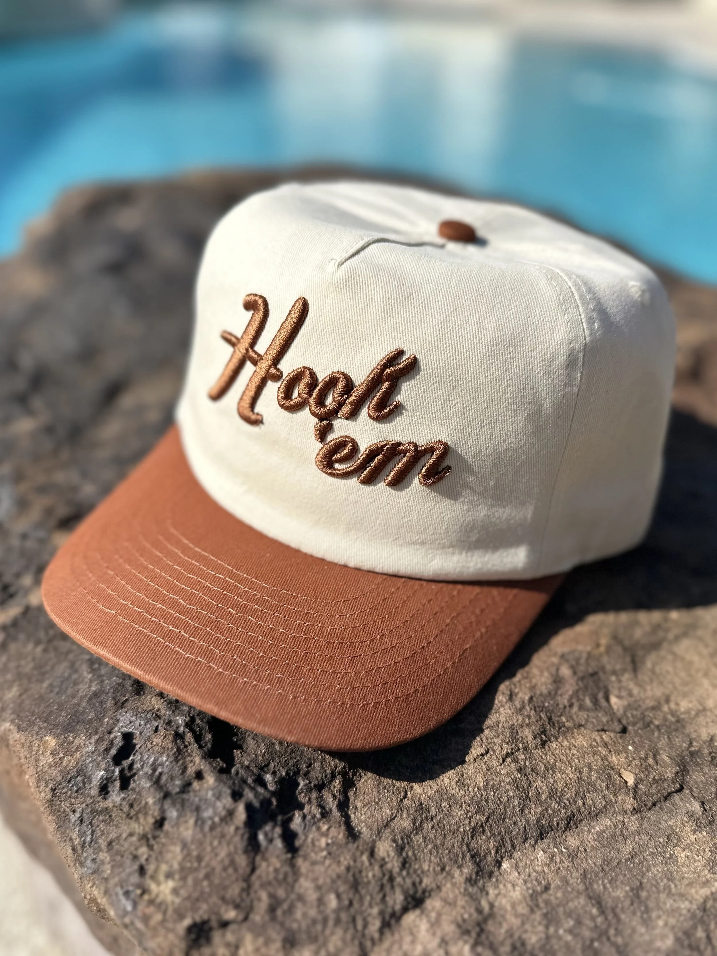 Cap with embroidery reading "Hoot Heme" resting on a rock by a swimming pool