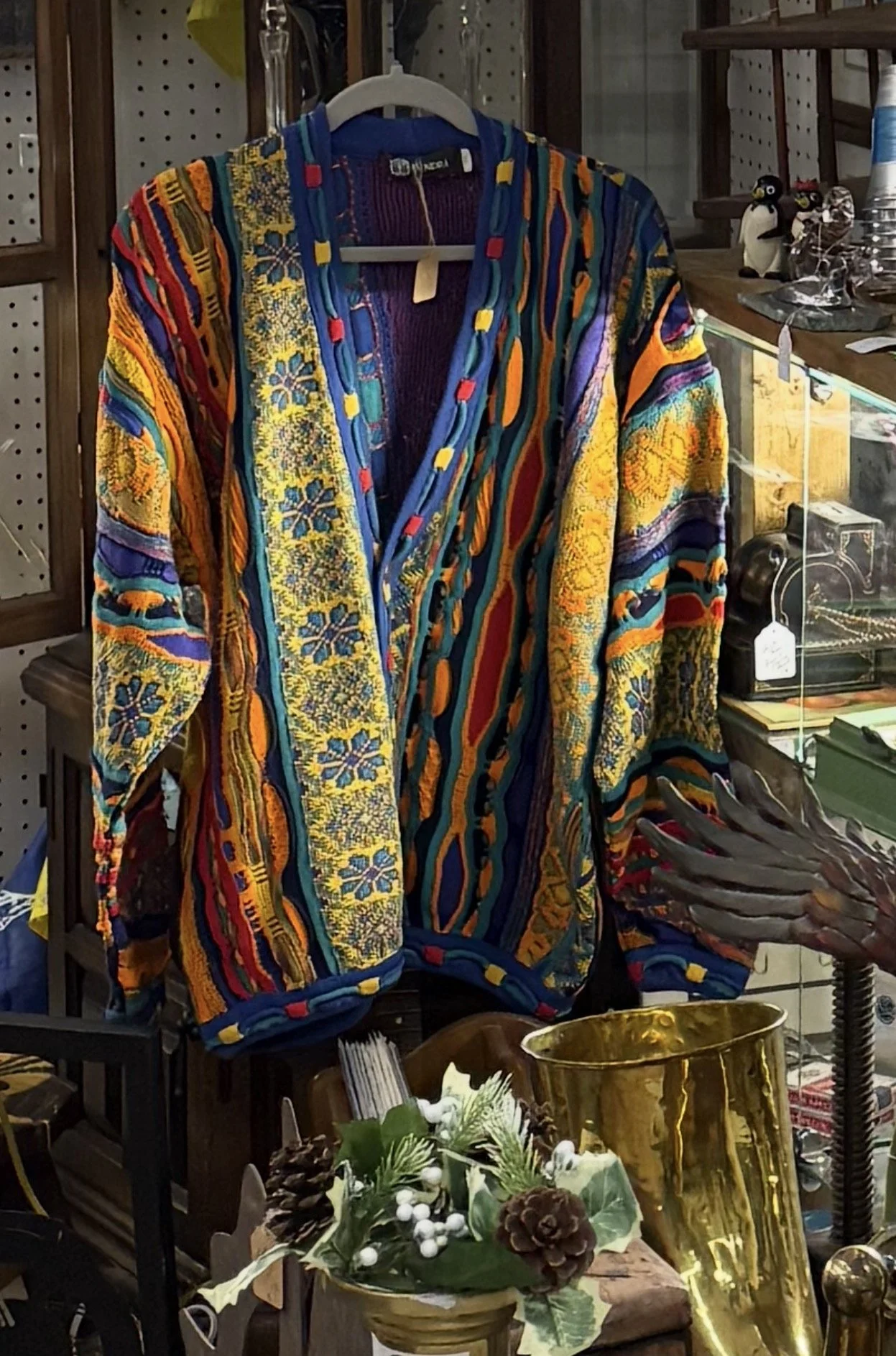 Colorful, embroidered jacket with intricate patterns hanging on a white hanger in an antique store with various decorative items and furniture around.