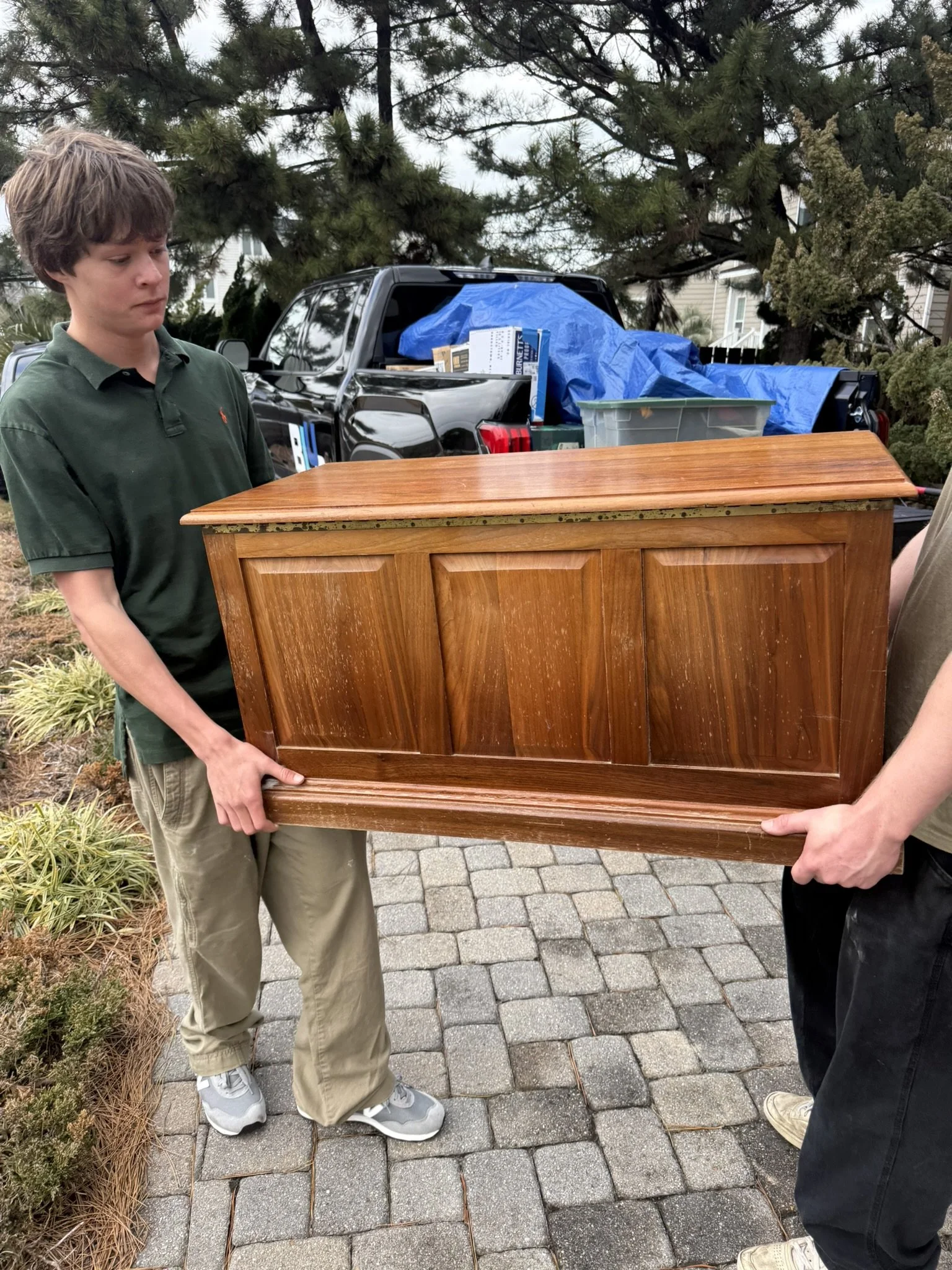 Two people are carrying a wooden piece of furniture, possibly a cabinet or dresser, outdoors on a paved pathway with greenery and a truck loaded with boxes and other items behind them.