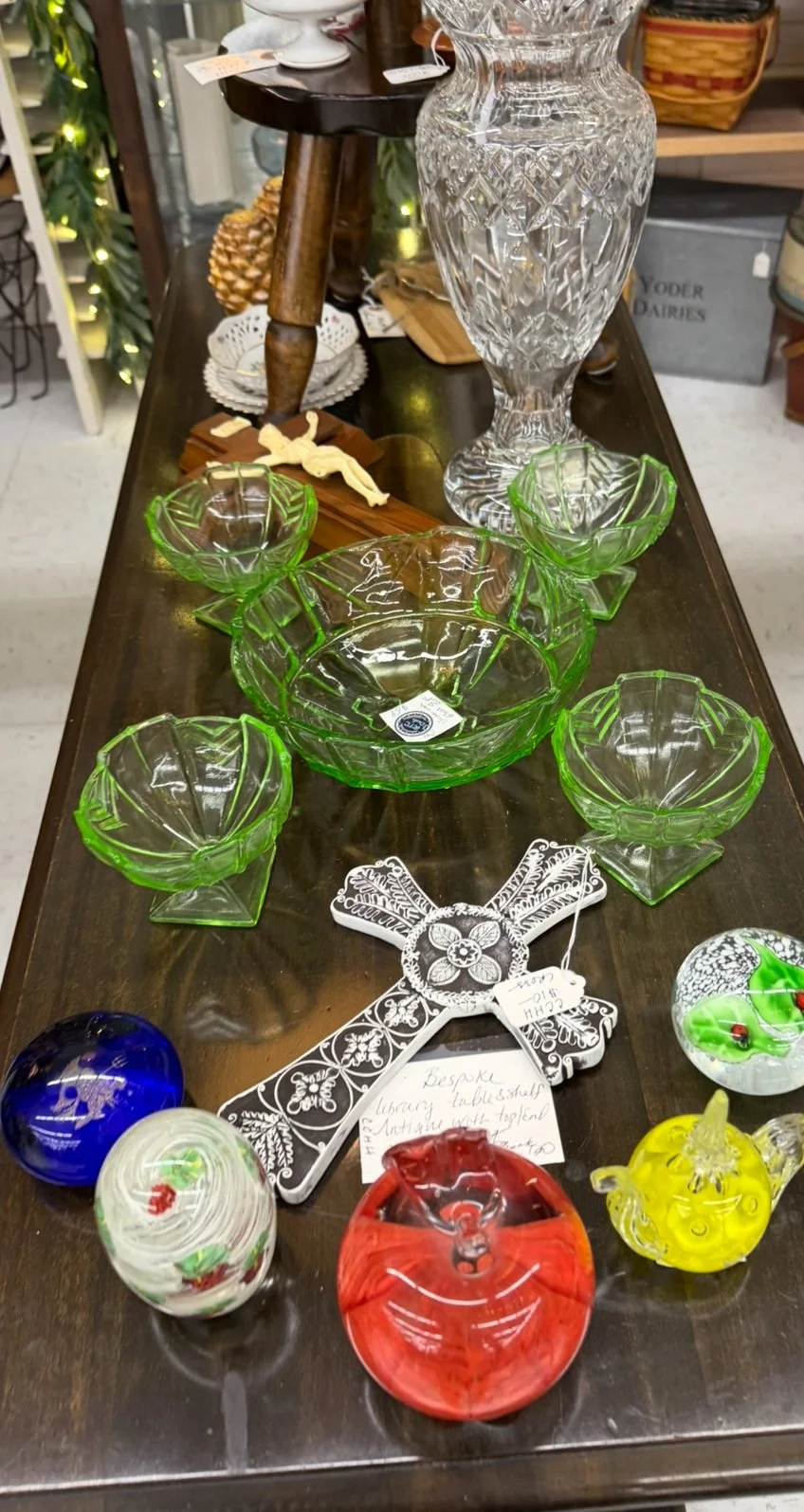 A variety of vintage glassware, including a large cut glass vase, green glass bowls, and colorful glass ornaments displayed on a wooden table.