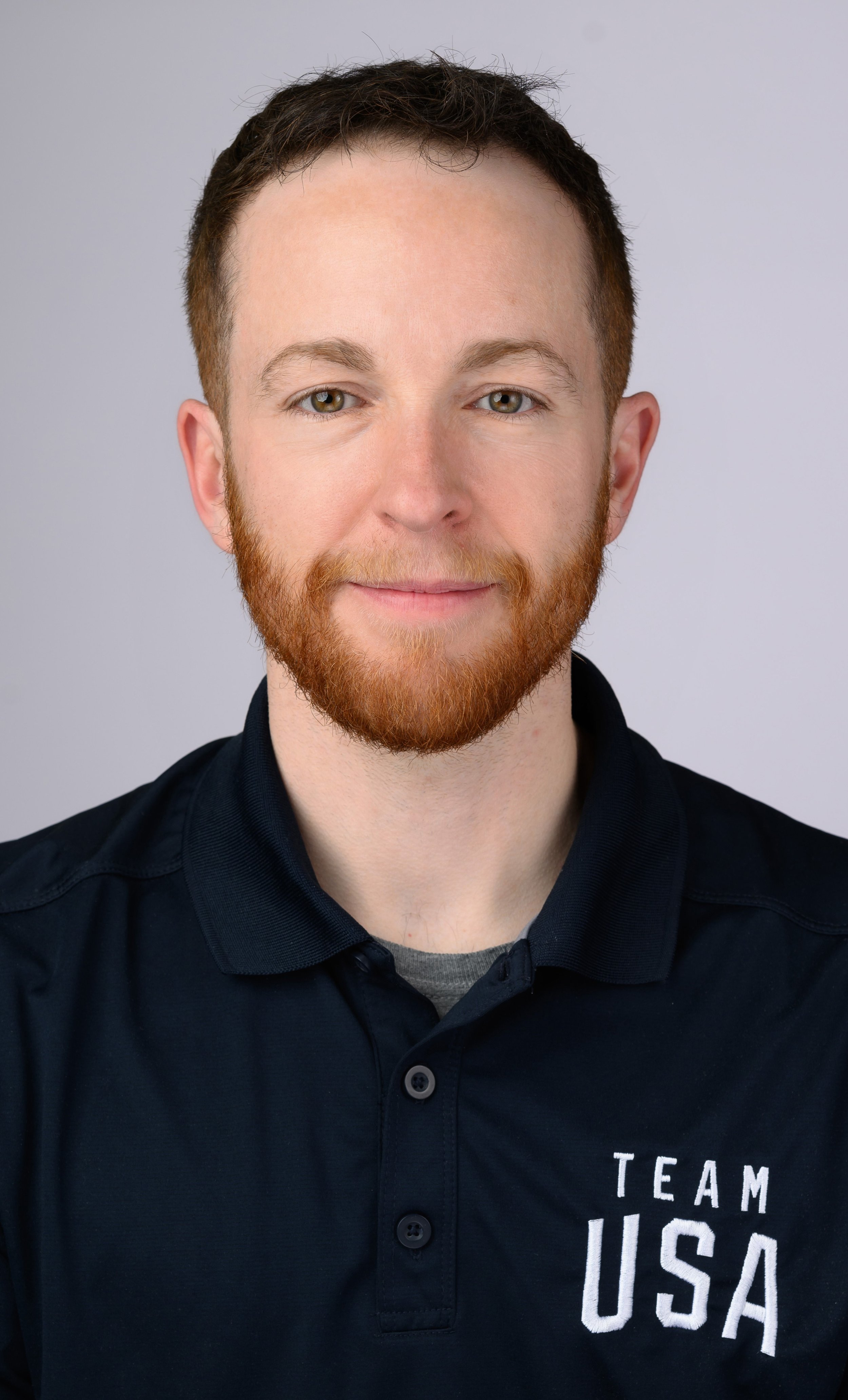 A young man with a beard and short brown hair, smiling, wearing a dark polo shirt with 'TEAM USA' text on it, against a light gray background.