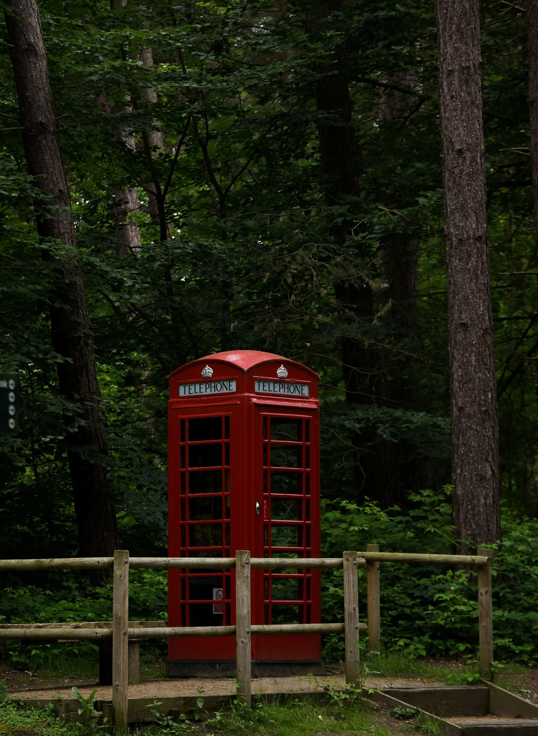 Red British-style telephone booth in a wooded area with green foliage, a wooden railing, and a dirt path.