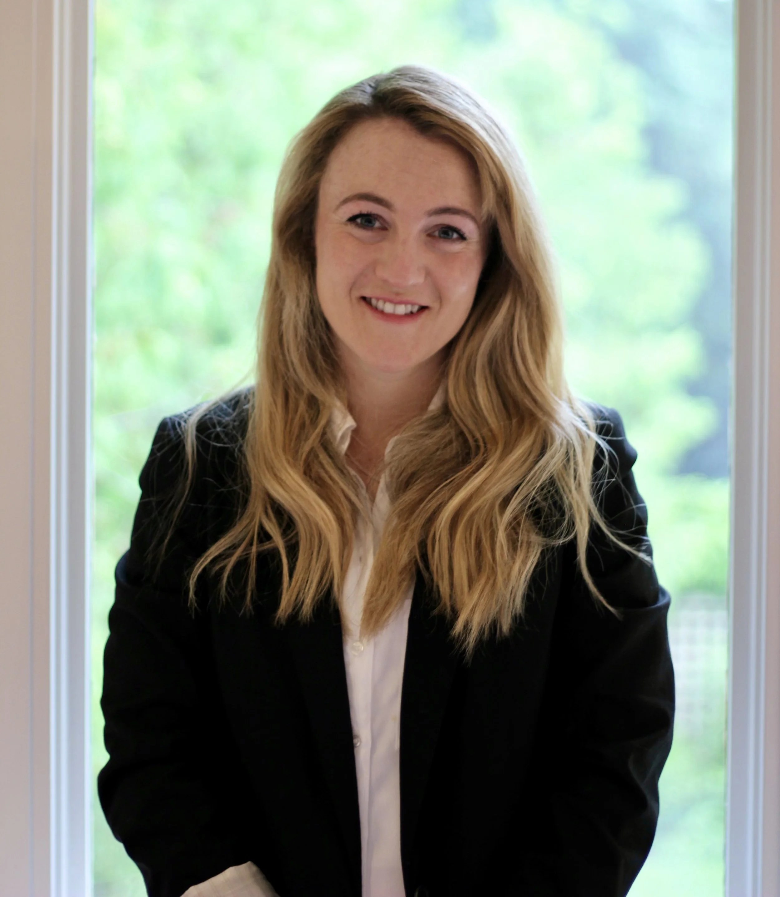 A woman with long, wavy blonde hair smiling in front of a window with greenery outside, wearing a black blazer and white shirt.
