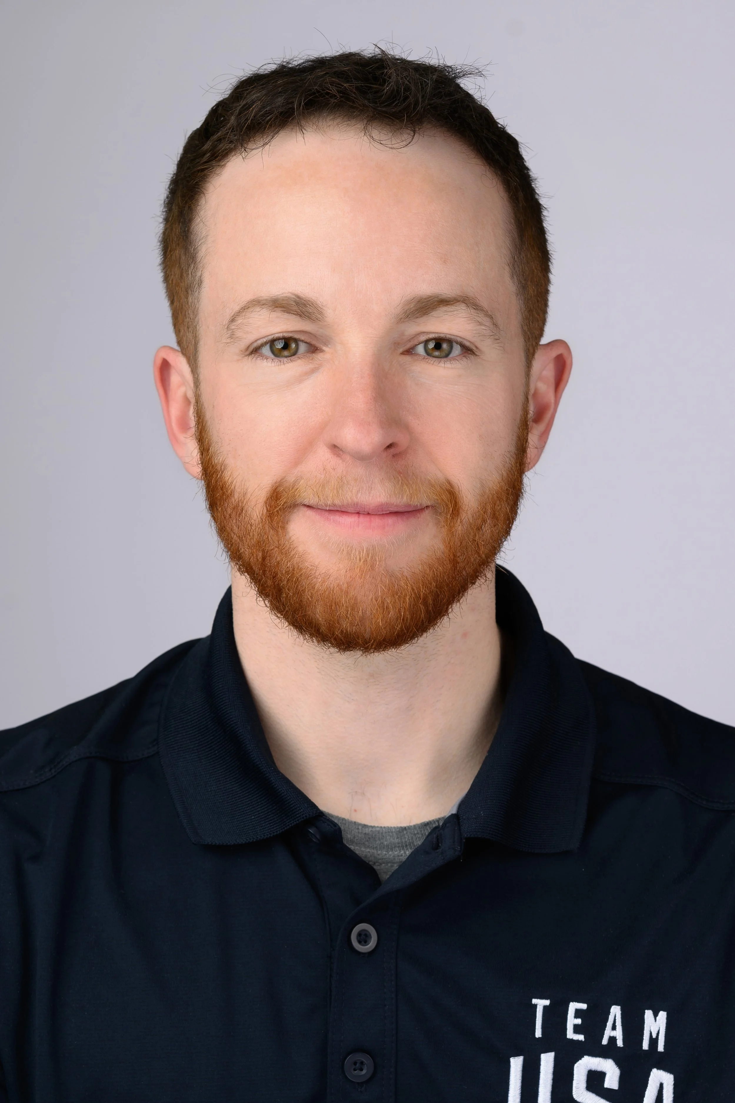 A young man with red hair and a beard, wearing a navy blue polo shirt with 'TEAM USA' written on it, smiling slightly at the camera.