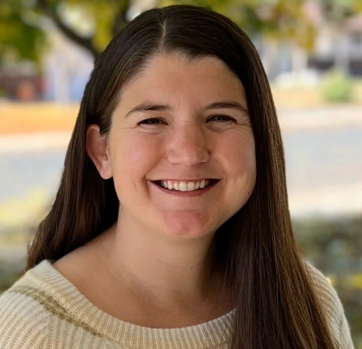 A woman with long brown hair smiling outdoors, wearing a cream-colored sweater.