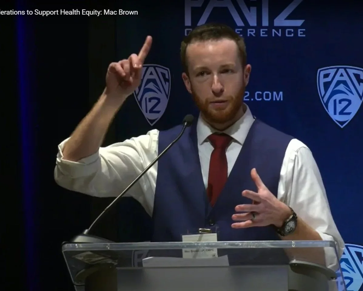 Man with red hair and beard in a suit vest and white shirt speaking at a podium with a PAC-12 conference backdrop, gesturing with one hand and pointing upwards with the other.
