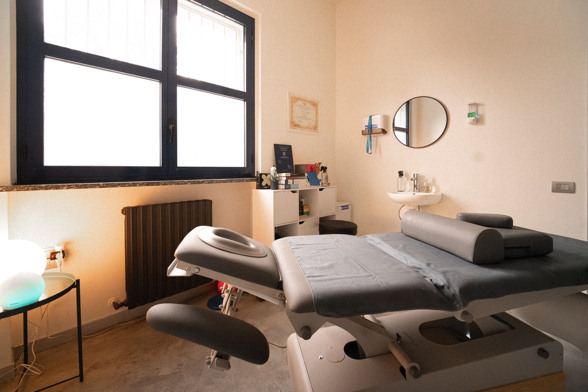 Una stanza di uno studio medico con un letto da esame, un piccolo lavabo e una finestra grande. La stanza è ben illuminata e ha arredi essenziali per consultazioni e trattamenti.