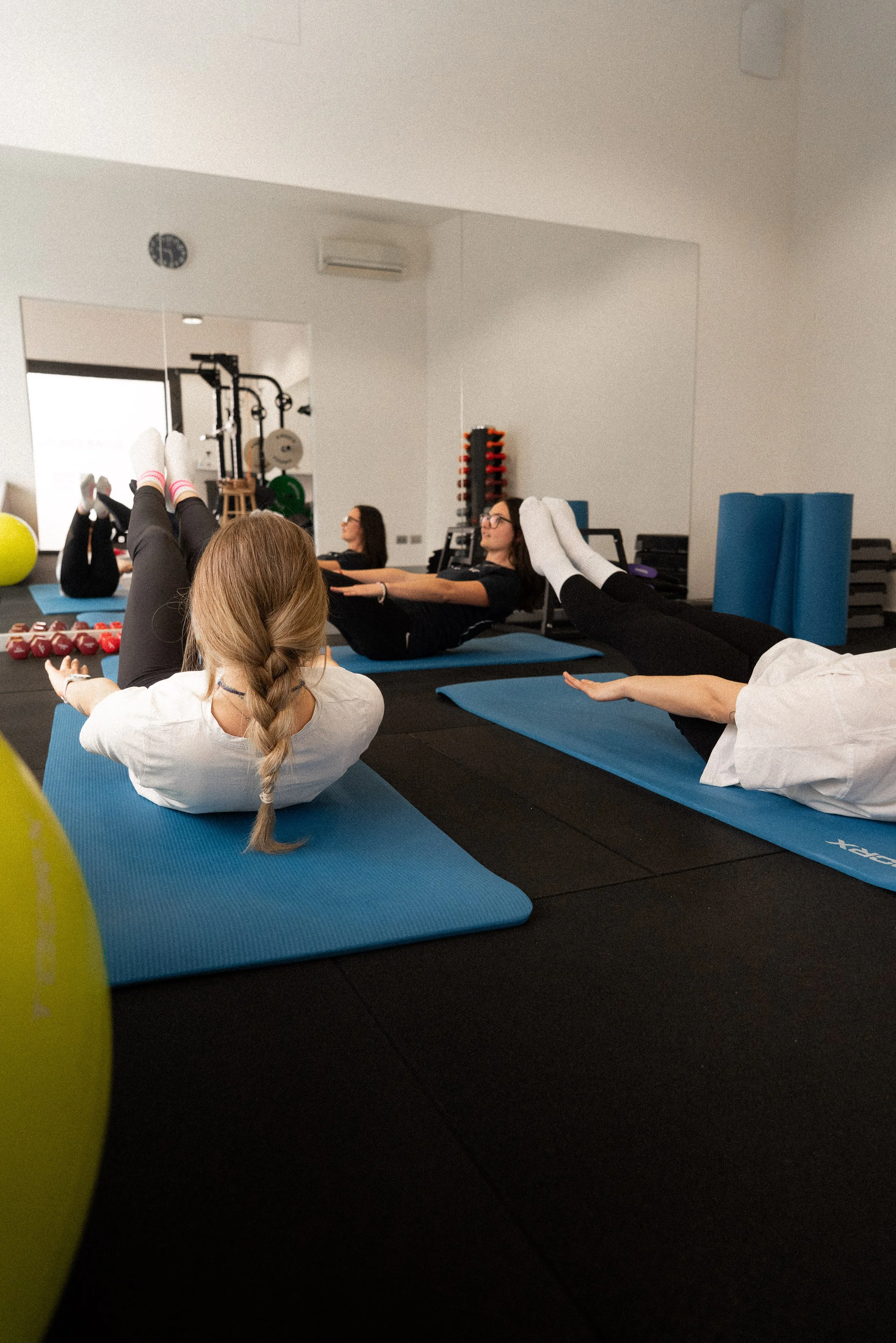 Persone che praticano yoga in una palestra con tappetini blu, in posizione di rilassamento, con attrezzi e attrezzature da palestra sullo sfondo.