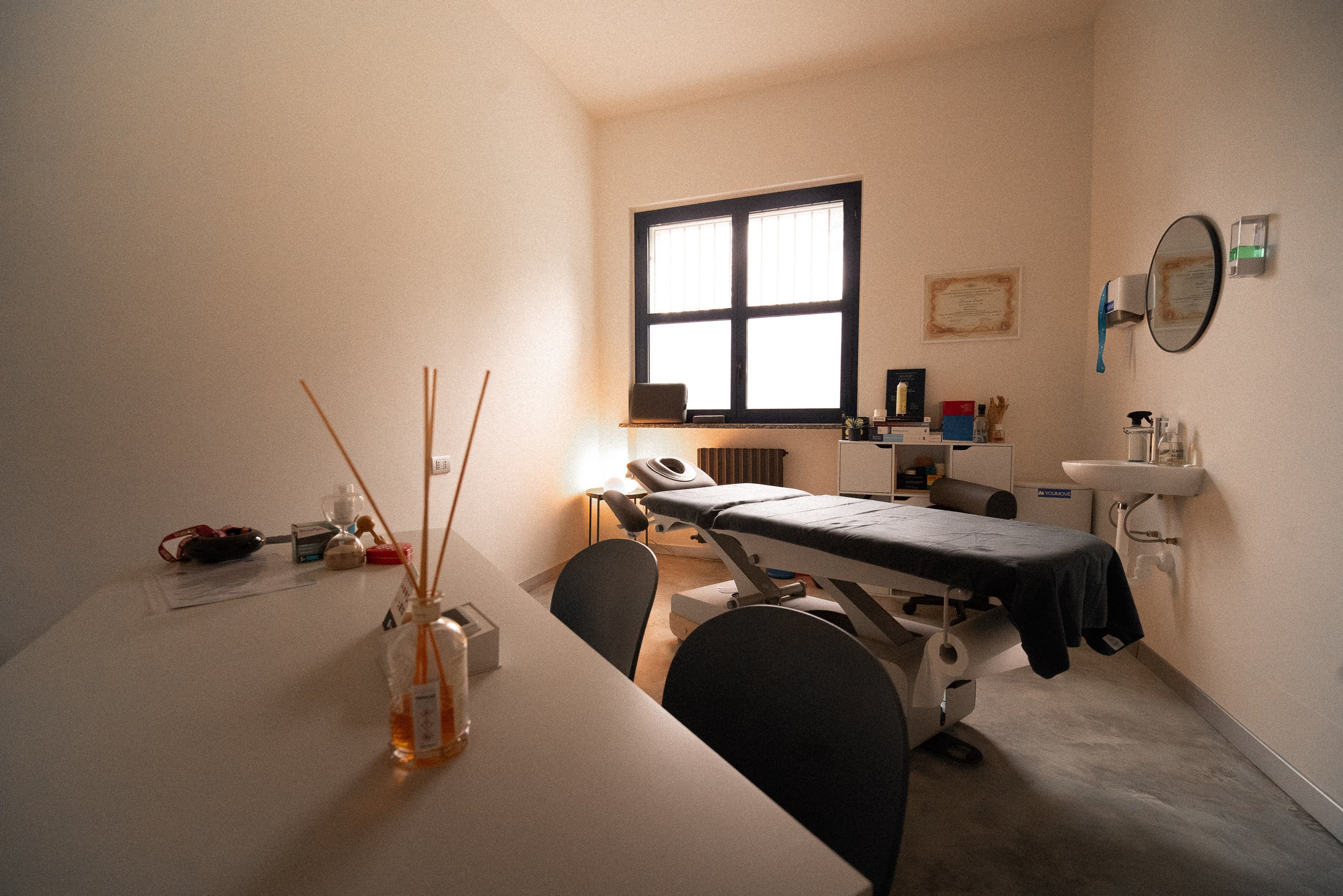 Ambiente di un studio medico con un lettino da visita, una finestra, una scrivania con oggetti e sedie, uno specchio e un lavabo.