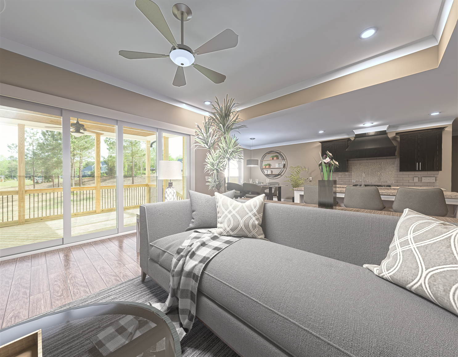 Modern living room with gray sofa, patterned pillows, and a blanket, open to a dining area and kitchen, with large sliding glass doors leading to a bright outdoor deck and green trees outside.