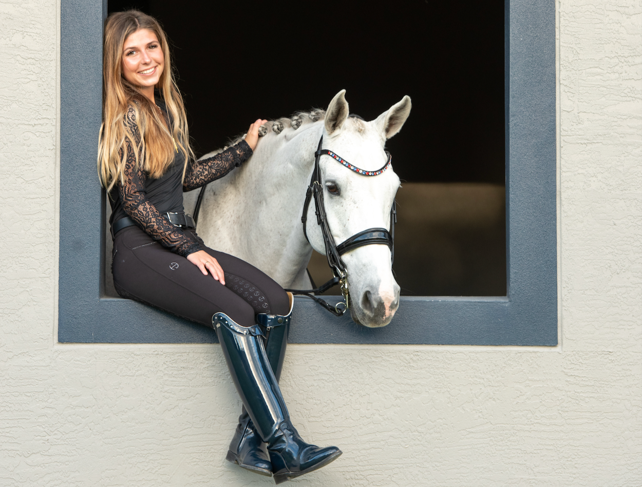A young woman with long blond hair and a black lace long sleeve top sitting on the window ledge of a stable, smiling at the camera, with her arm resting on a white horse that is looking out of the window.