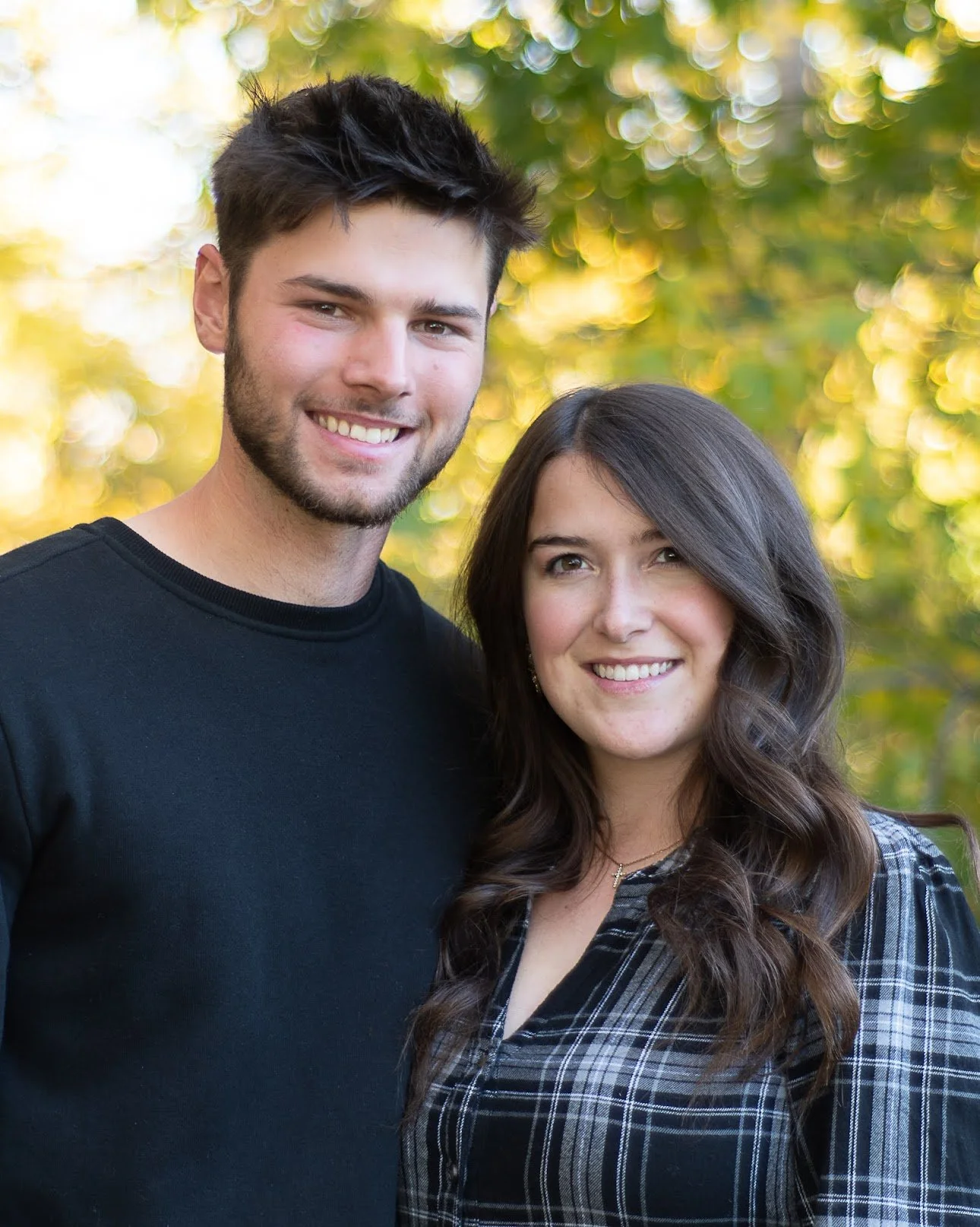 A young man and woman smiling outdoors with trees and yellow sunlight in the background.