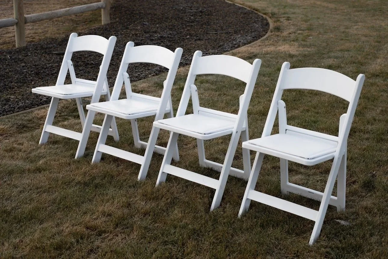 Four white folding chairs arranged in a row on a grassy outdoor area near a flower bed.