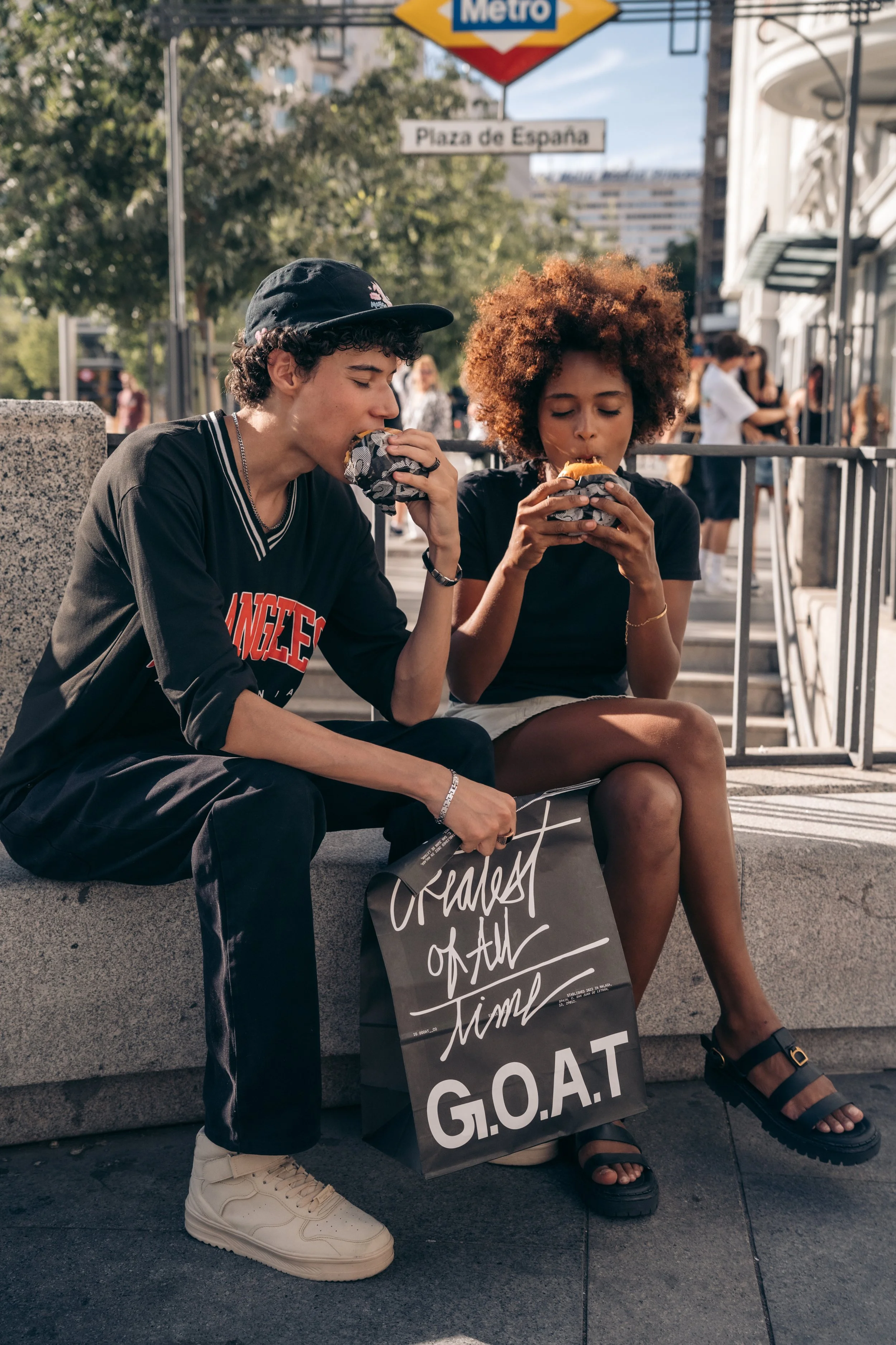 Jóvenes comiendo hamburguesas en la calle, con un cartel que dice 'Greatest of All Time' y un letrero de metro y plaza de España en el fondo.