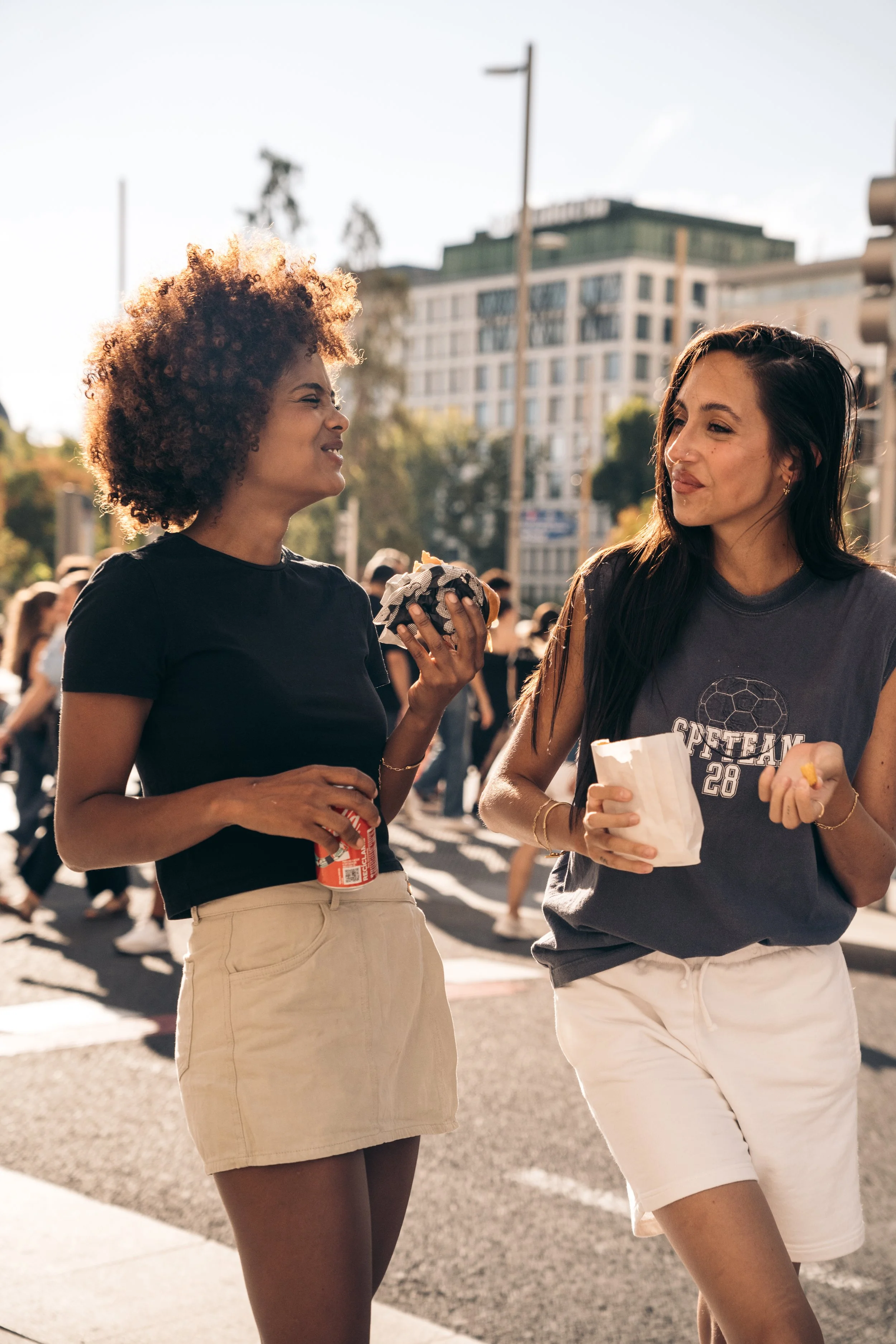 Dos mujeres jóvenes caminando por la ciudad, una con un sándwich y una con una lata de soda, parecen estar conversando y disfrutando del día.