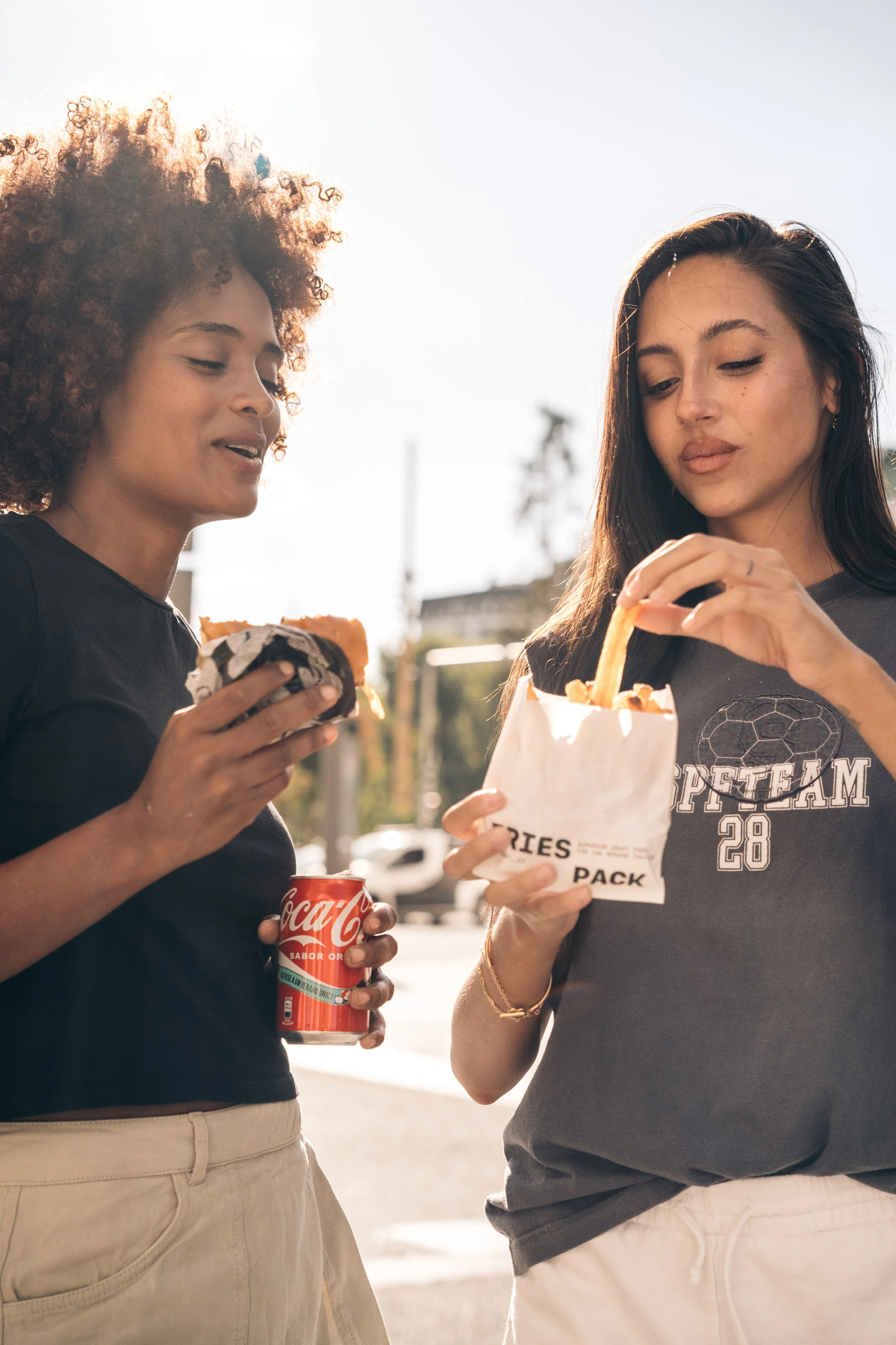 Dos mujeres jóvenes disfrutando comida rápida al aire libre, con una enciendo en un día soleado.