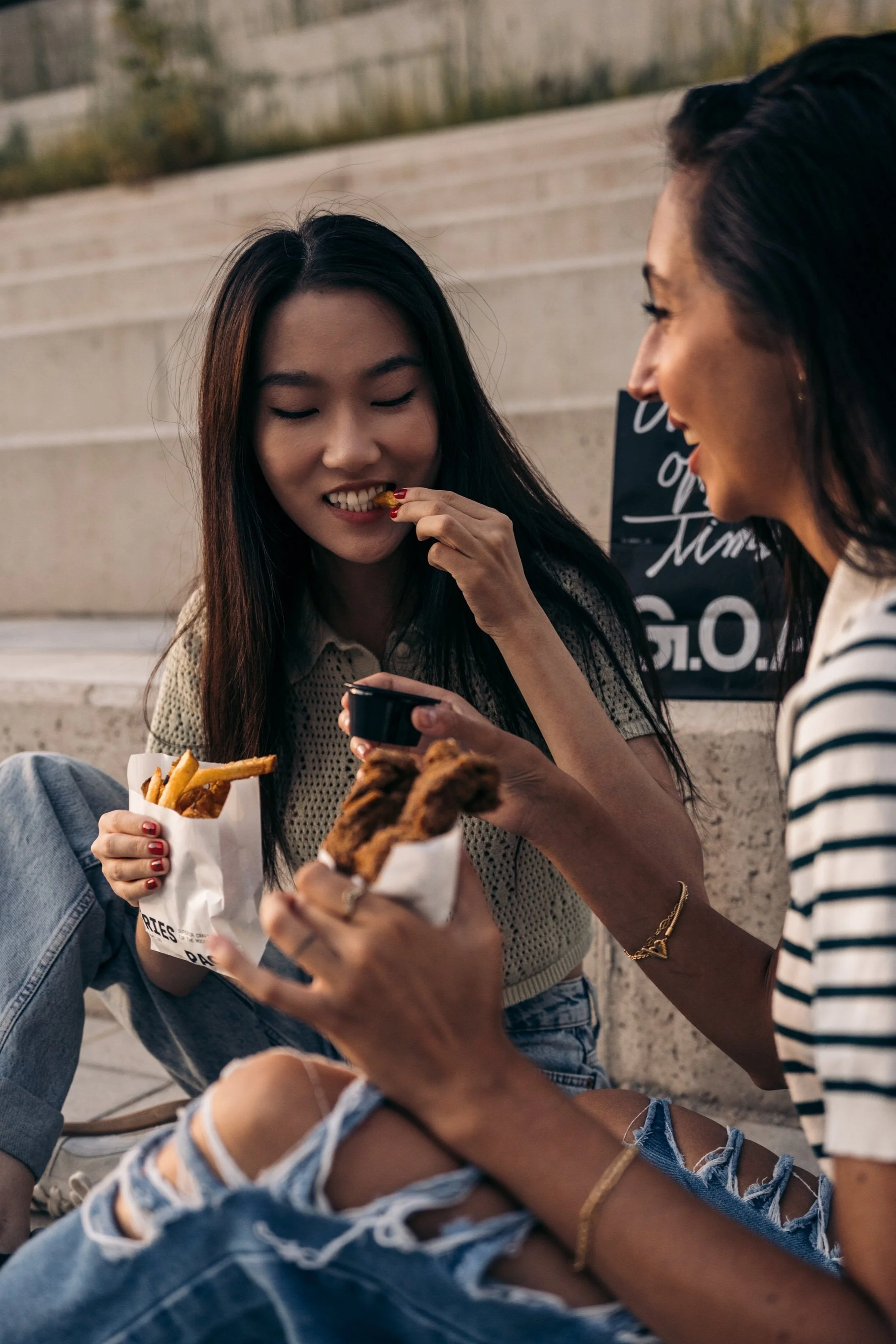 Dos mujeres sentadas en las escaleras disfrutando comida rápida, una con papas fritas y otra con pollo frito, compartiendo un momento juntas.
