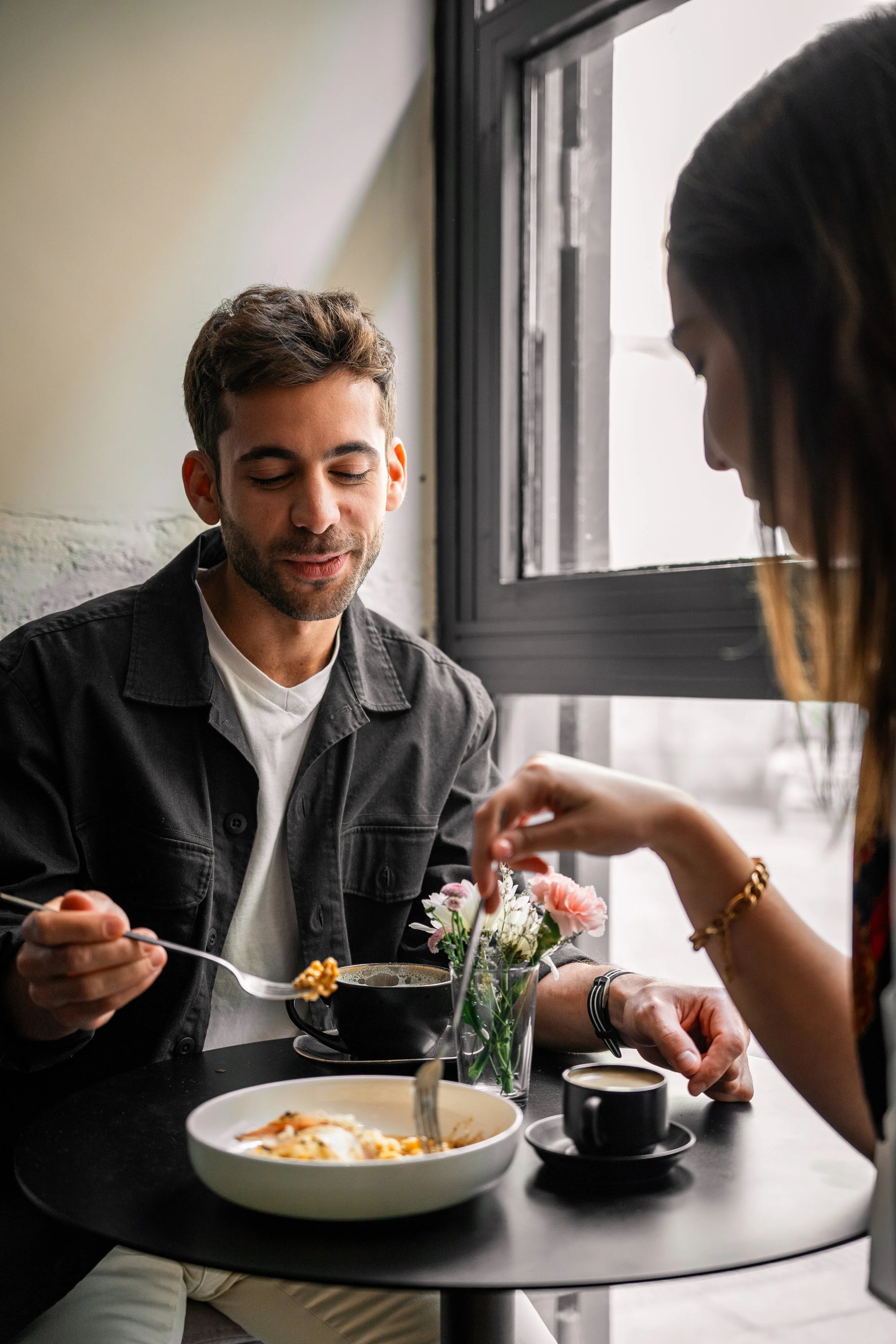 Una pareja sentada en una mesa de café, con comida y bebidas, compartiendo un momento en un lugar con mucha luz natural y decoración moderna.
