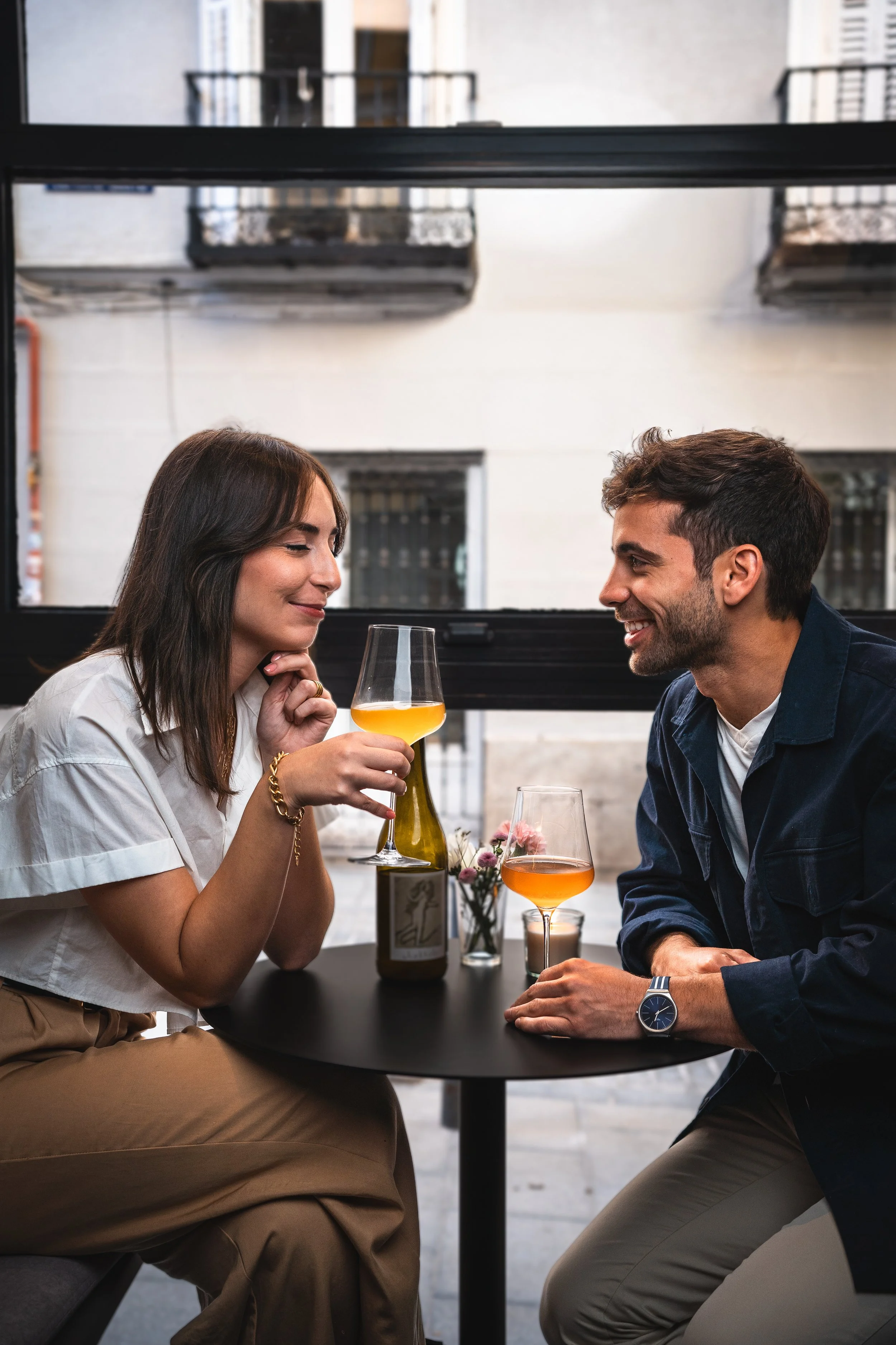 Una pareja sentada en una mesa de café, brindando con copas de vino rosado, en un ambiente moderno y acogedor.