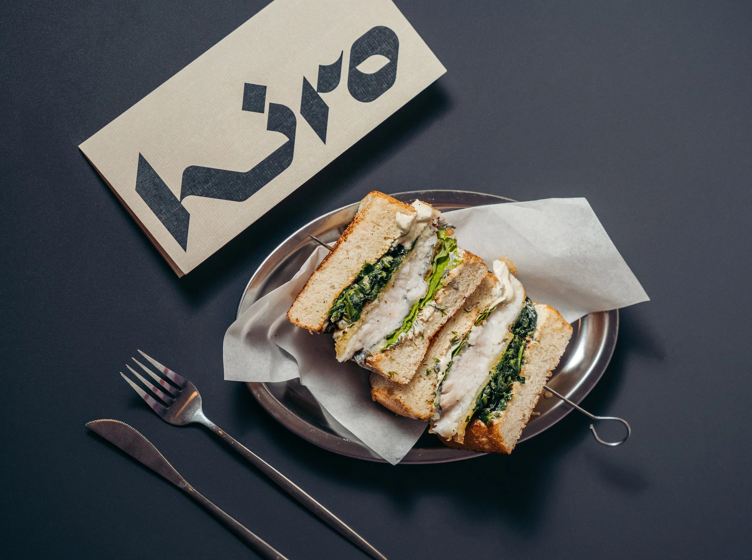 Sándwich en un plato con hojas verdes y queso, con un tenedor y cuchillo a su lado, sobre una mesa oscura y un cartel que dice 'LUN'.