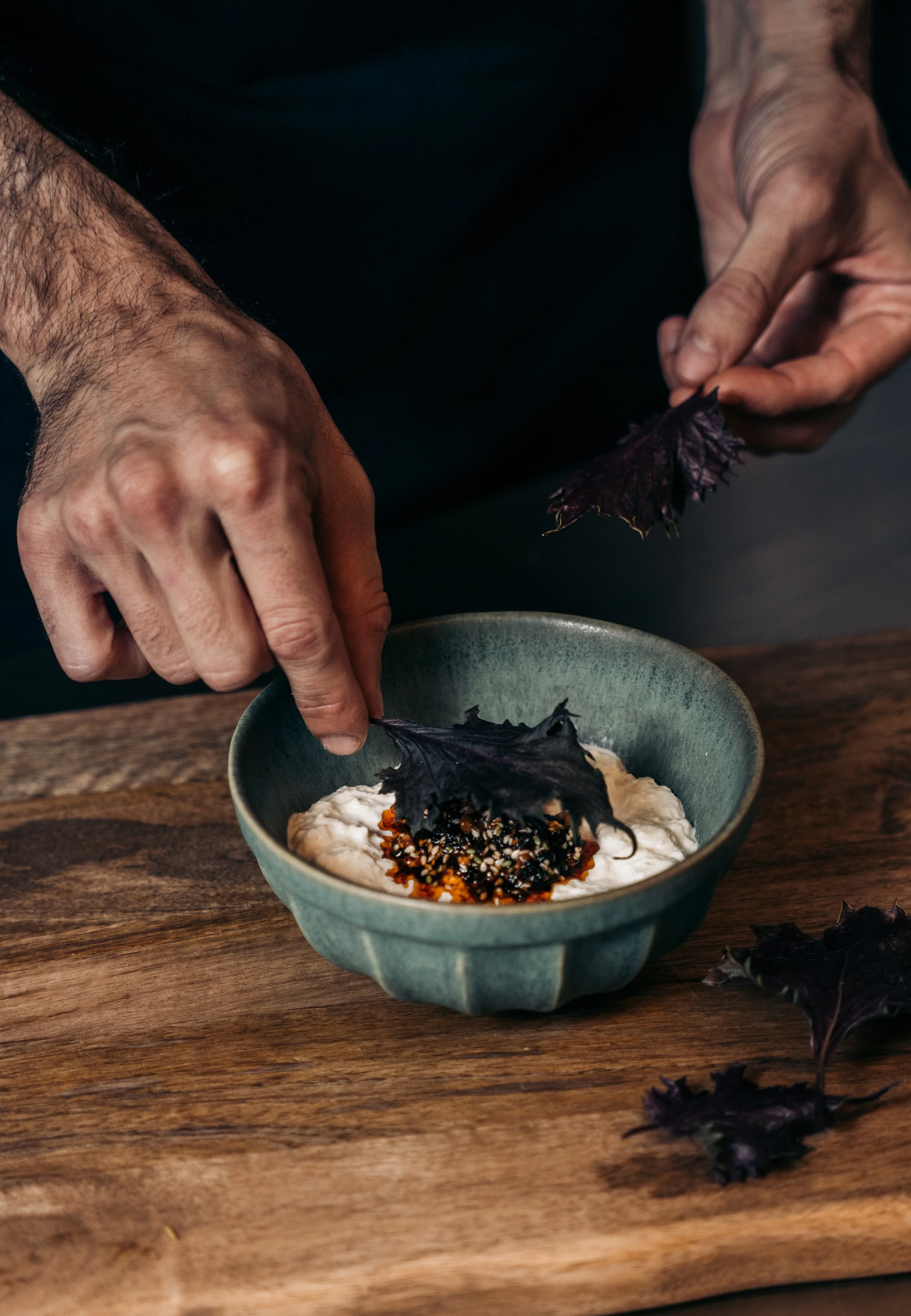Mano colocando una hoja de capucha negra sobre un plato con sopa en un tatami de madera.