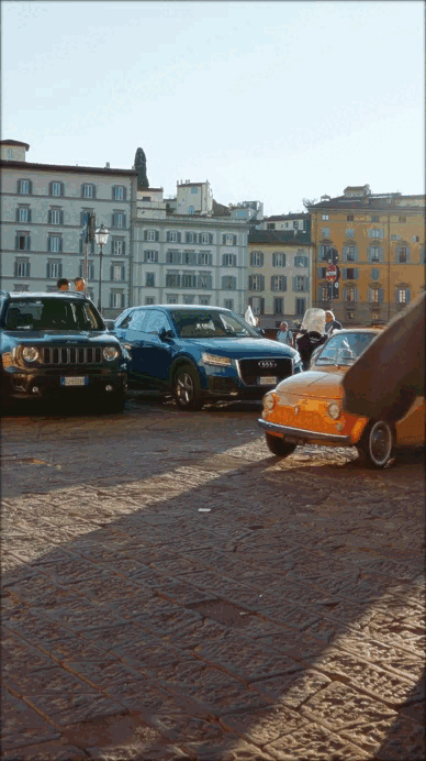 Estacionamiento con varios autos en una plaza urbana, con edificios históricos al fondo y el suelo de piedra.