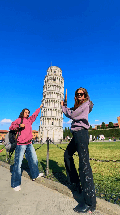 Dos mujeres frente a la Torre de Pisa tocándola y sonriendo, en un día soleado con cielo azul.