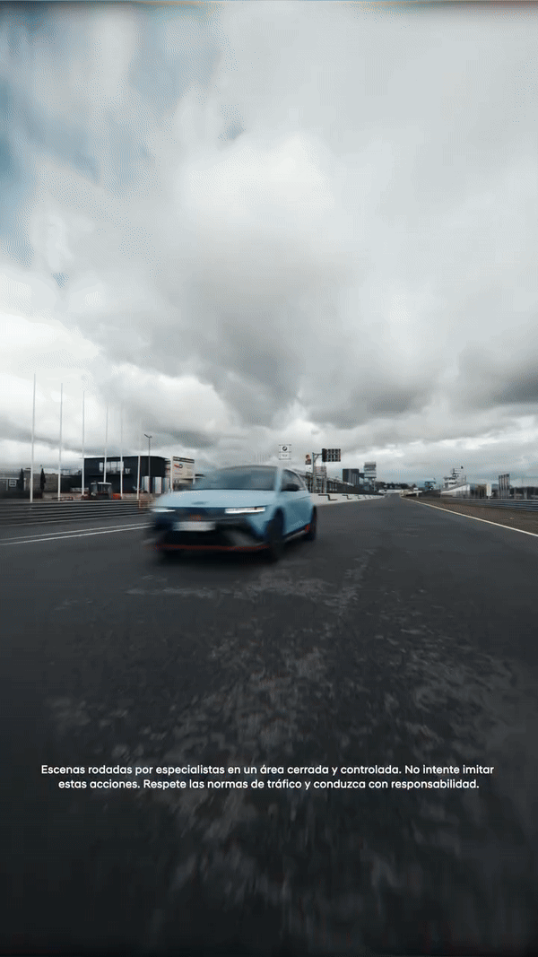 Un automóvil azul en movimiento en una pista de carrera cubierta por nubes grises, con edificios y señalización al fondo.
