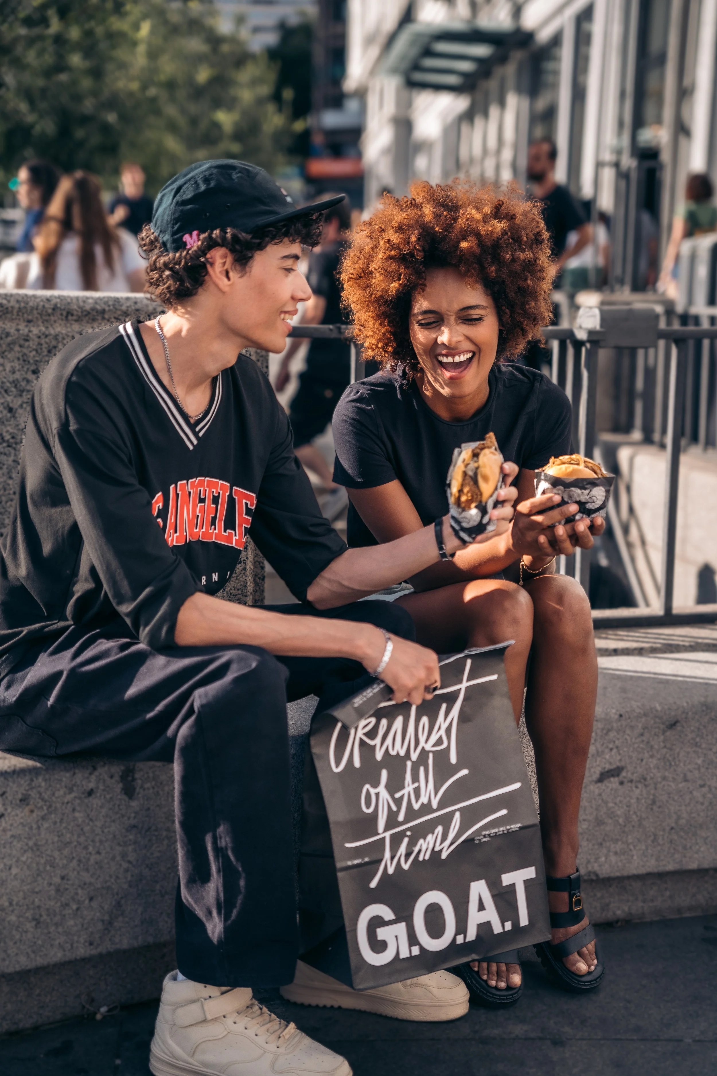 Dos personas sentadas en una banca riendo y compartiendo hamburguesas en una calle concurrida