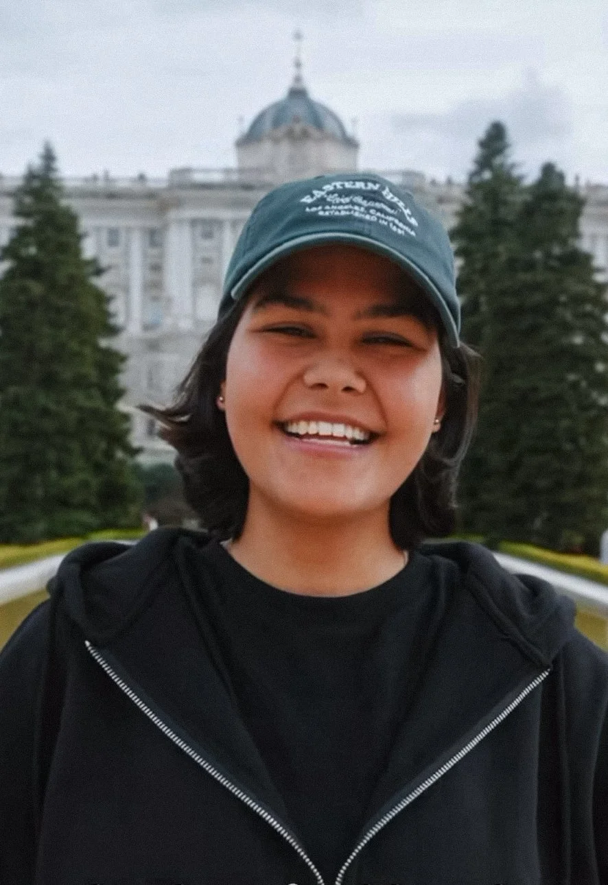 Persona sonriendo en exteriores, con edificio histórico y árboles en el fondo.