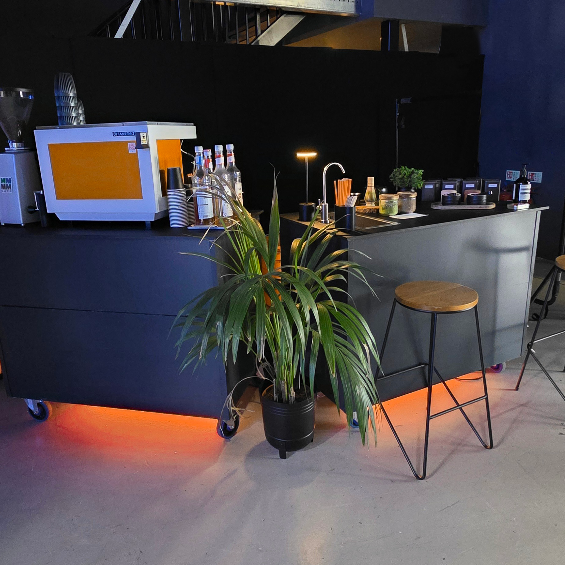 A black bar counter with a potted plant in front, bar stools on the side, and various beverage supplies on the counter, illuminated by orange accent lighting and small lamp.
