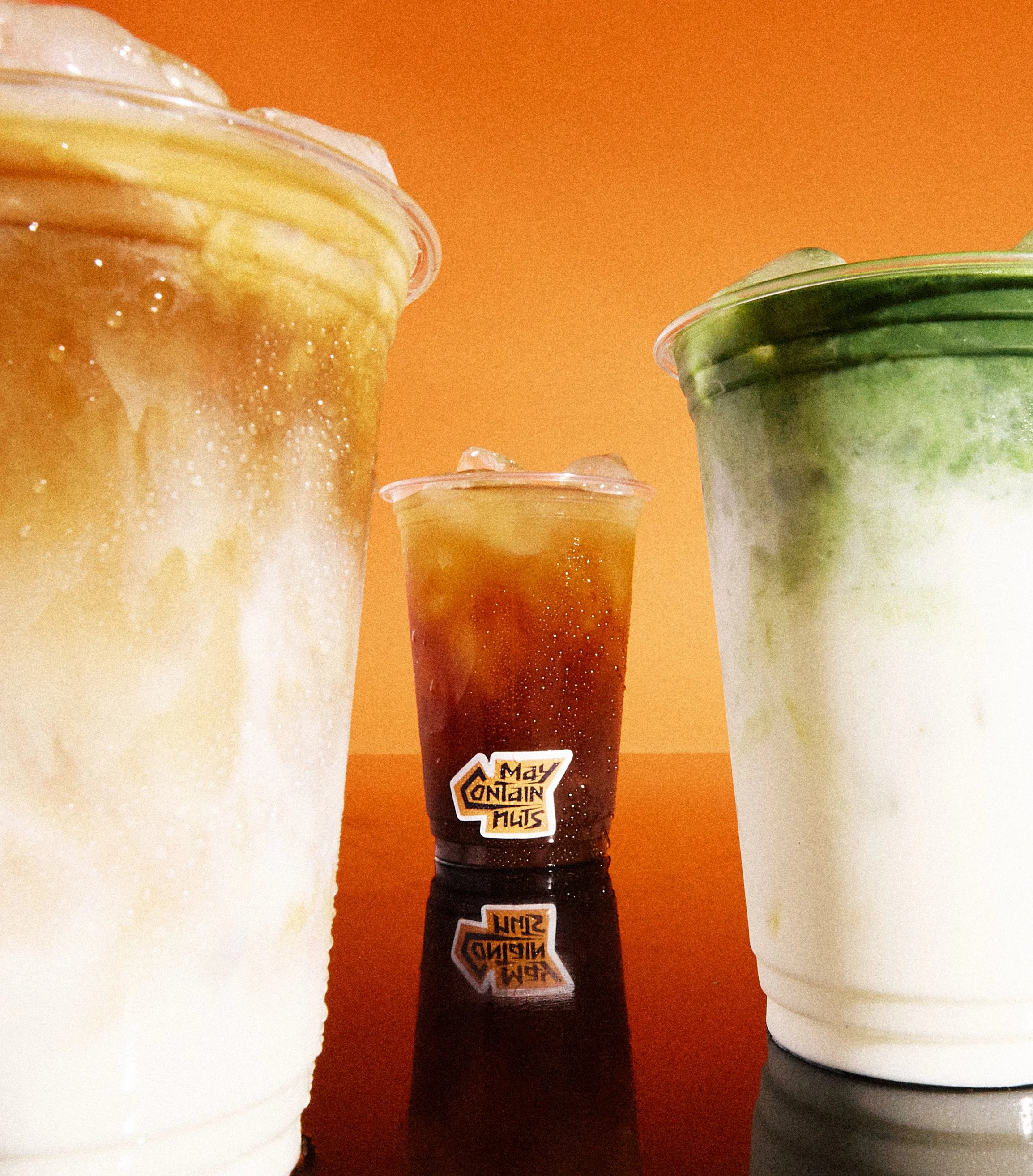 Three cold beverages in clear plastic cups with condensation, set against an orange background. The drinks appear to be iced coffee or tea and matcha latte.