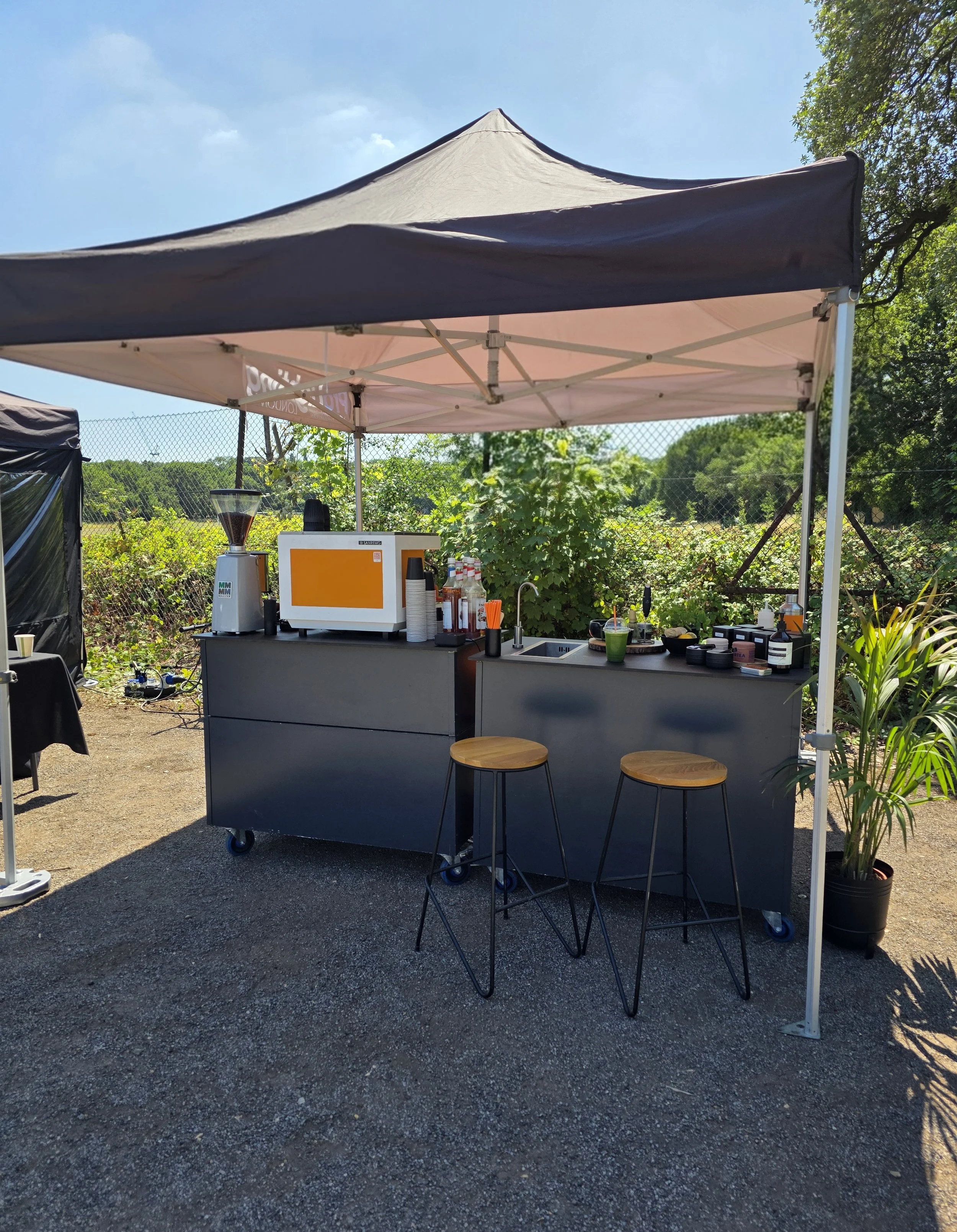An outdoor coffee stand under a beige canopy with a gray counter, bar stools, and a coffee grinder on the left, surrounded by greenery and a chain-link fence.