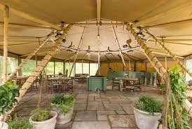 Inside a large tent with wooden support beams and tables, with potted plants around the entrance.