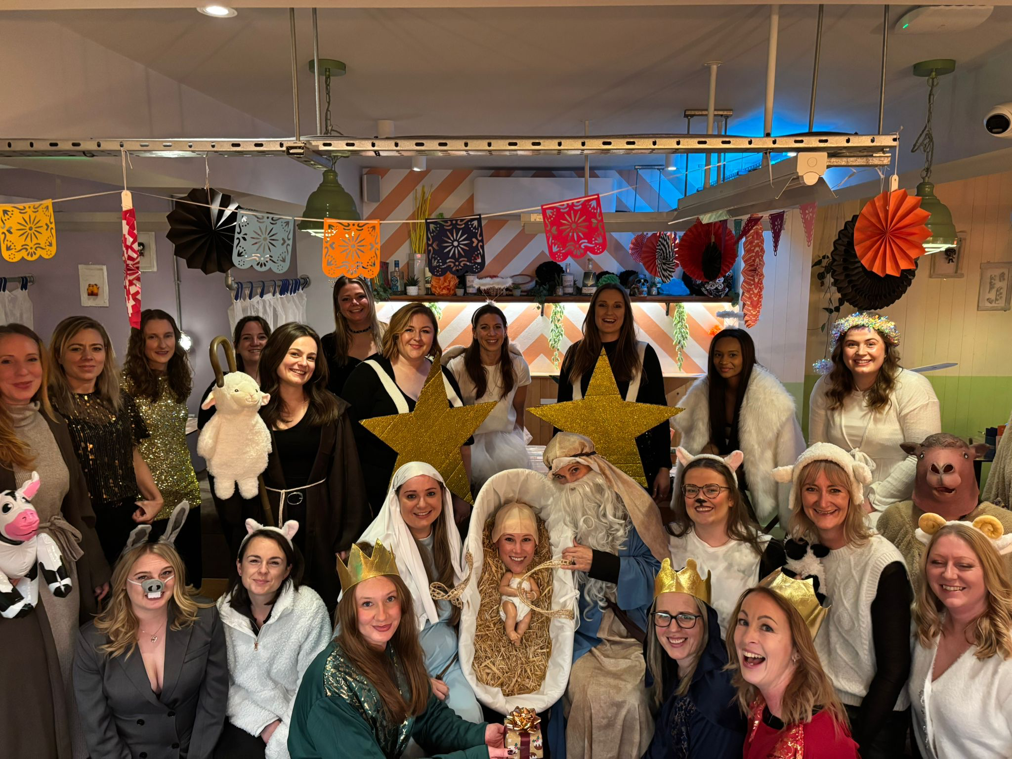 Group of women at festive party dressed in costumes including animals, angels, kings, and other characters; decorated with colorful banners, paper fans, hanging garlands, and fairy lights.