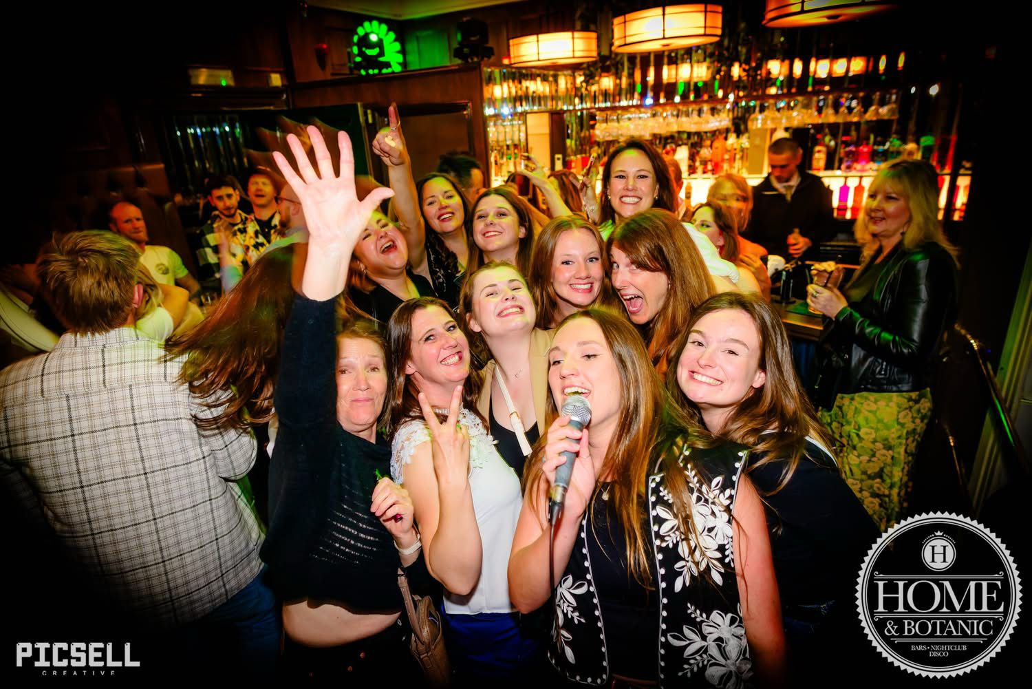 Group of women laughing and singing with a microphone at a bar or nightclub, lively atmosphere with colorful lights and a bar in the background.