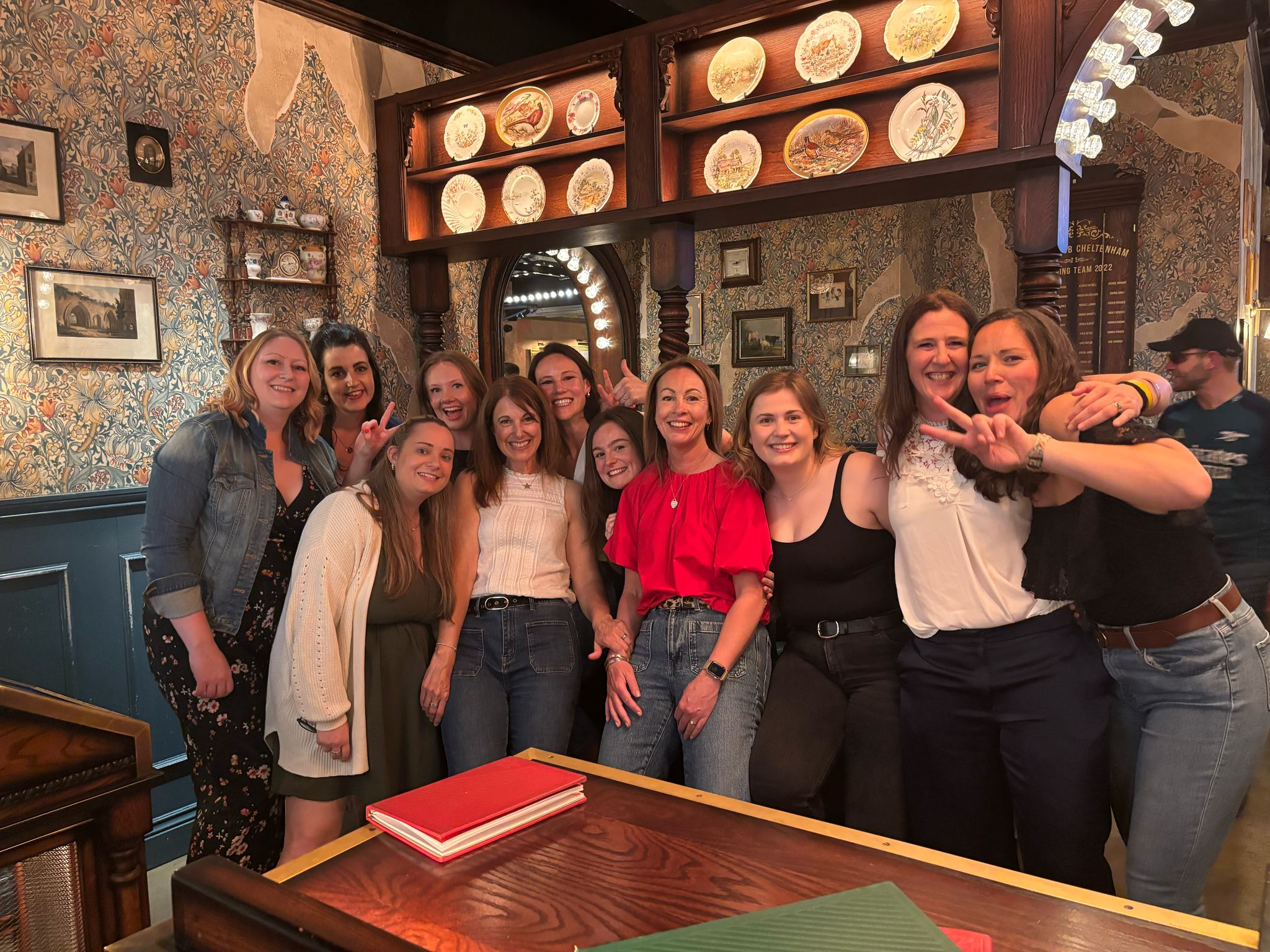 A group of eleven women smiling and posing together in a restaurant with patterned wallpaper and decorative plates on shelves.
