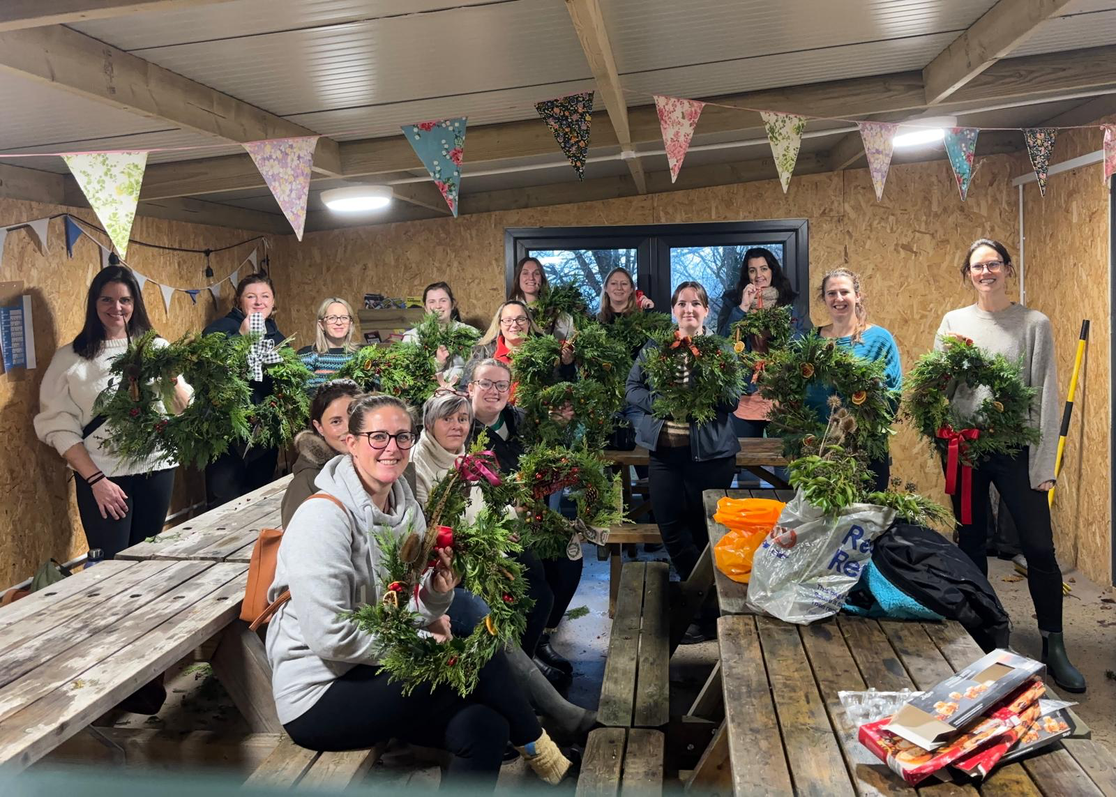 Group of people inside a wood-paneled room, decorating Christmas wreaths with greenery, ornaments, and ribbons, with bunting hanging from the ceiling.