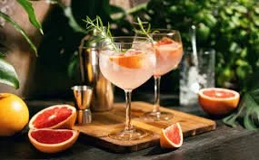 Two pink cocktails with fruit garnishes on a wooden board, surrounded by grapefruit halves and other citrus fruits outdoors.
