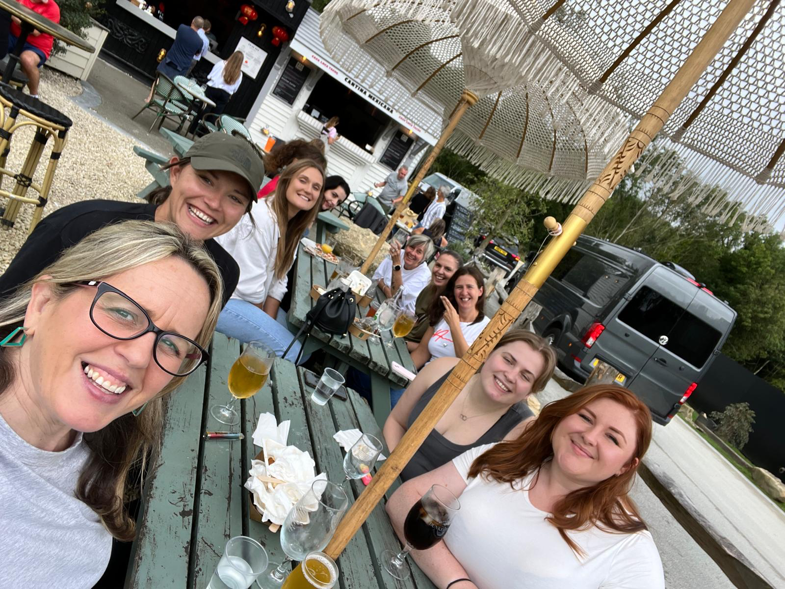A group of nine women sitting and standing around a long outdoor table at a restaurant, smiling, with drinks and food in front of them, under large umbrellas.